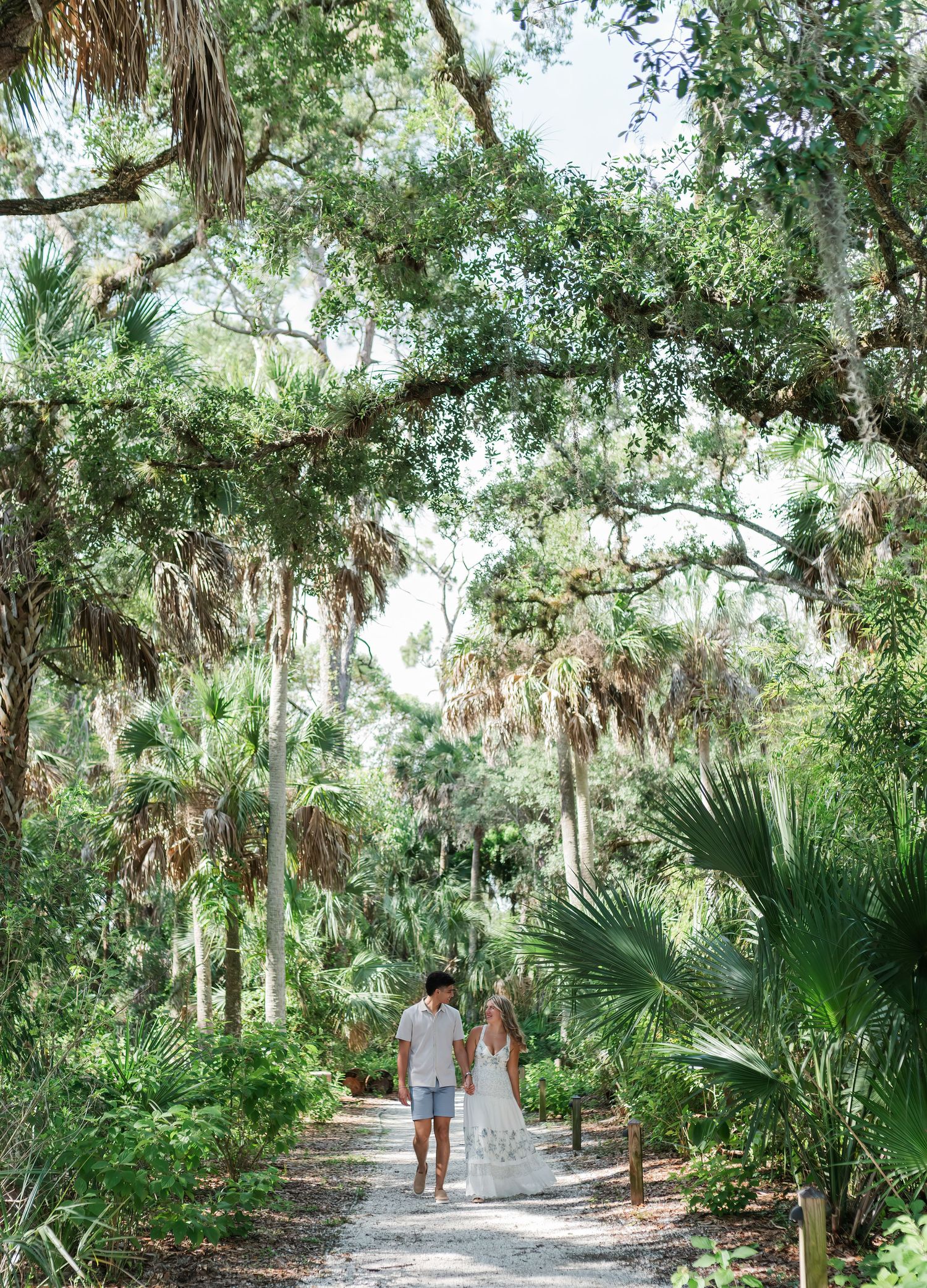 A couple walks hand in hand along a lush, tree-lined path surrounded by tropical Florida greenery.
