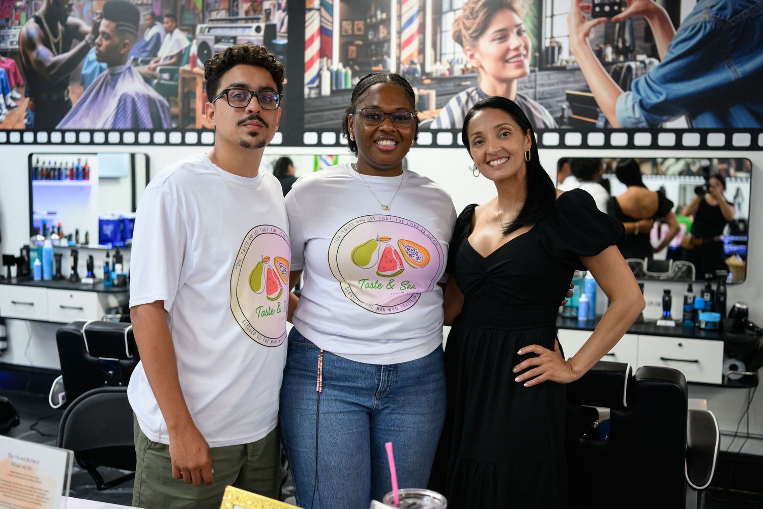 Three people posing together in a barbershop, two wearing matching white logo t-shirts, smiling at the camera.