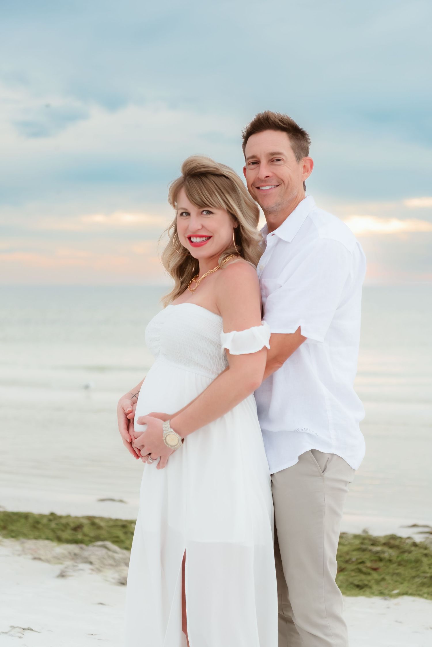 Pregnant couple in white outfits embracing on a beach at sunset, maternity photo session.