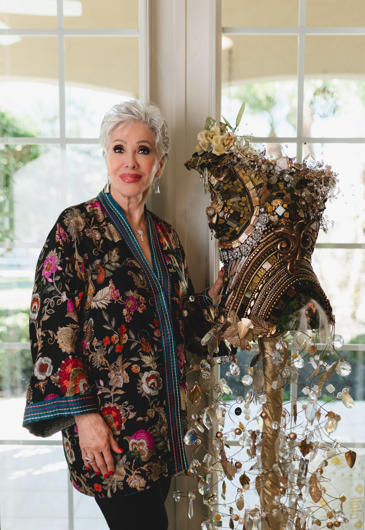 Elegant woman with white hair in floral jacket poses beside ornate jeweled corset display near sunlit windows.