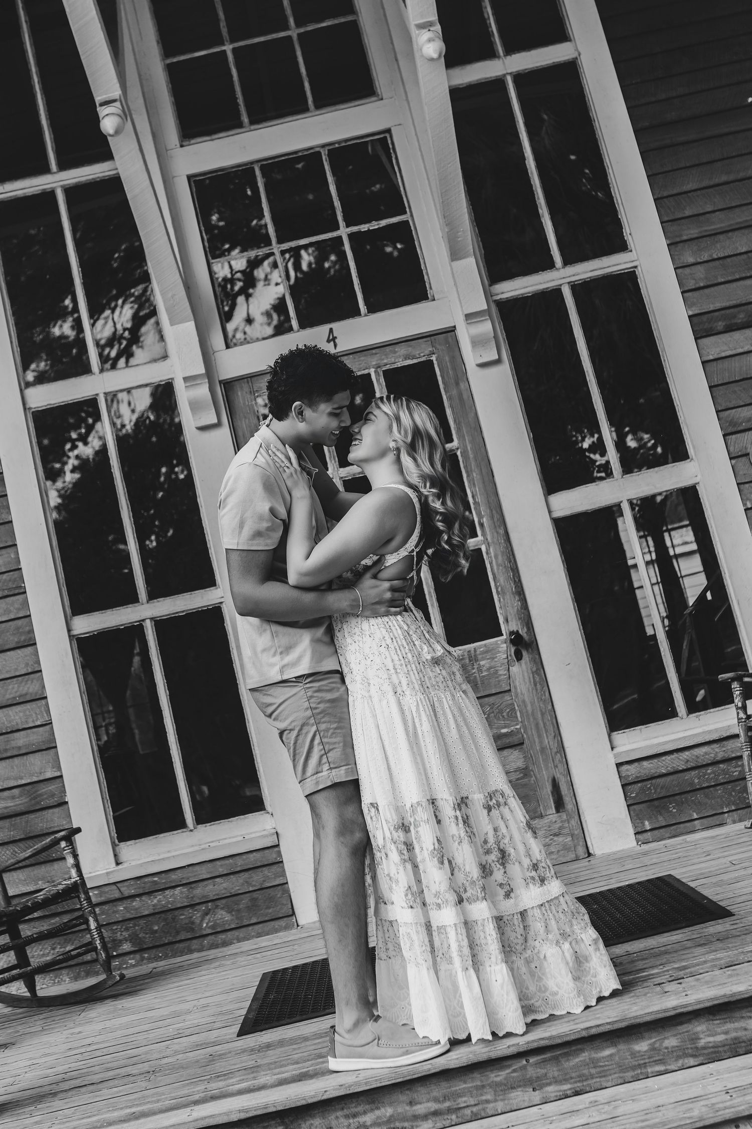 Black and white photo of a couple sharing a kiss on a wooden porch in front of large French doors.