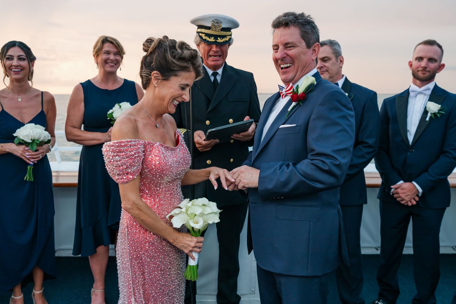 Couple exchanging rings at outdoor wedding ceremony on ship deck with bridal party in navy dresses at sunset.