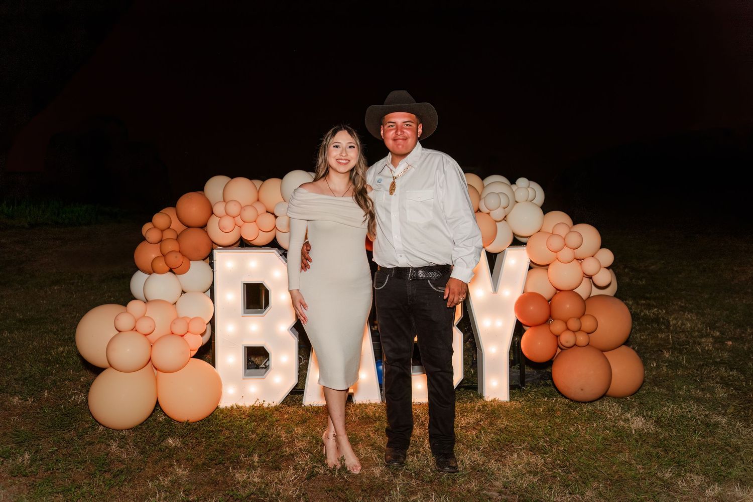 A couple stands in front of a balloon arch with illuminated 'BOY' letters at an outdoor gender reveal party.
