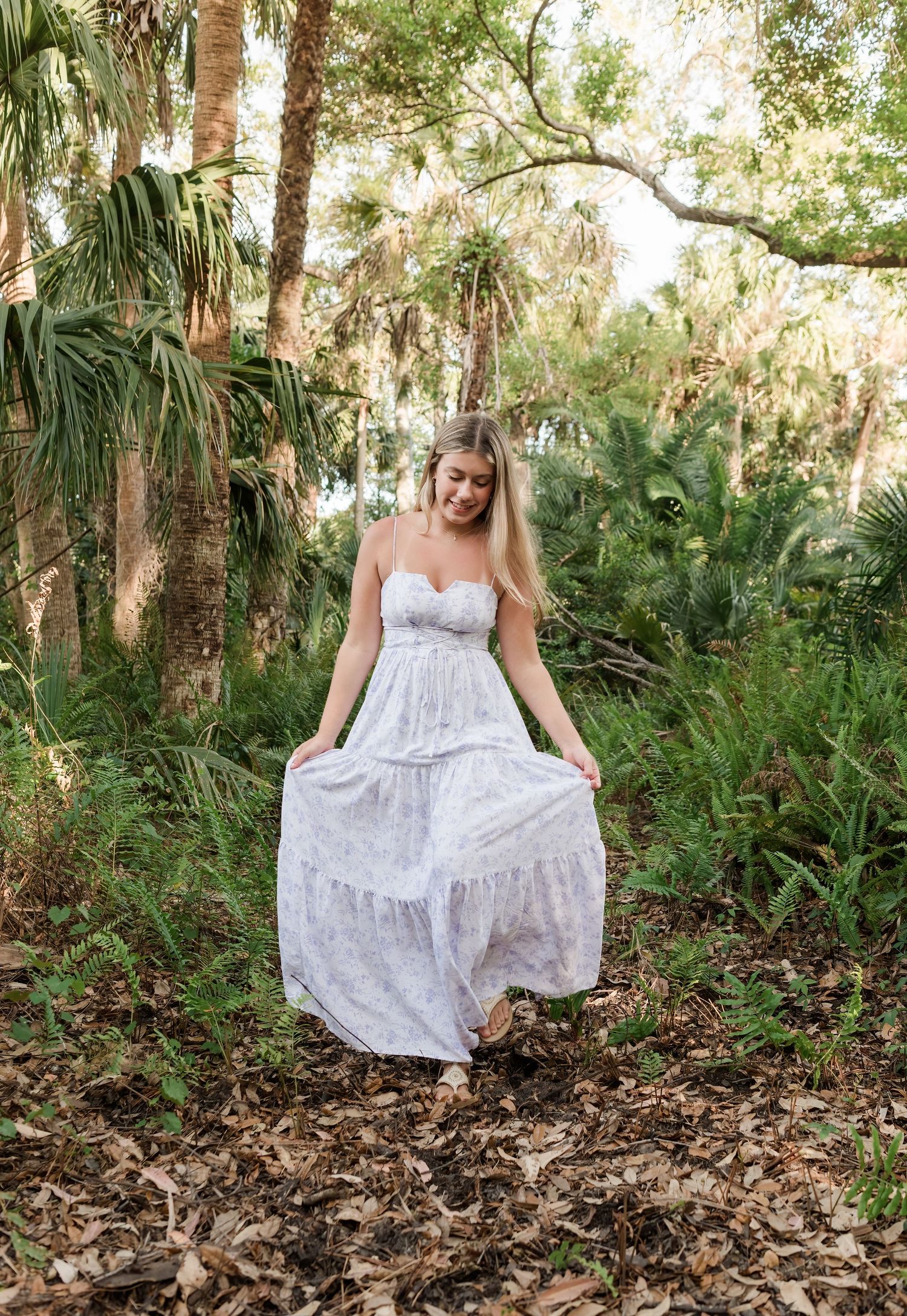 A person in a flowing white lace maxi dress stands on fallen leaves in a lush tropical forest setting.