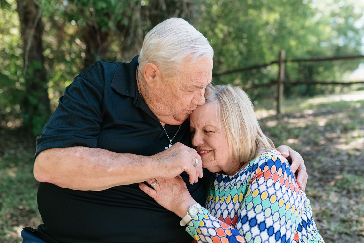 Elderly couple sharing a tender moment outdoors, man kissing woman's head while holding hands near a wooden fence.