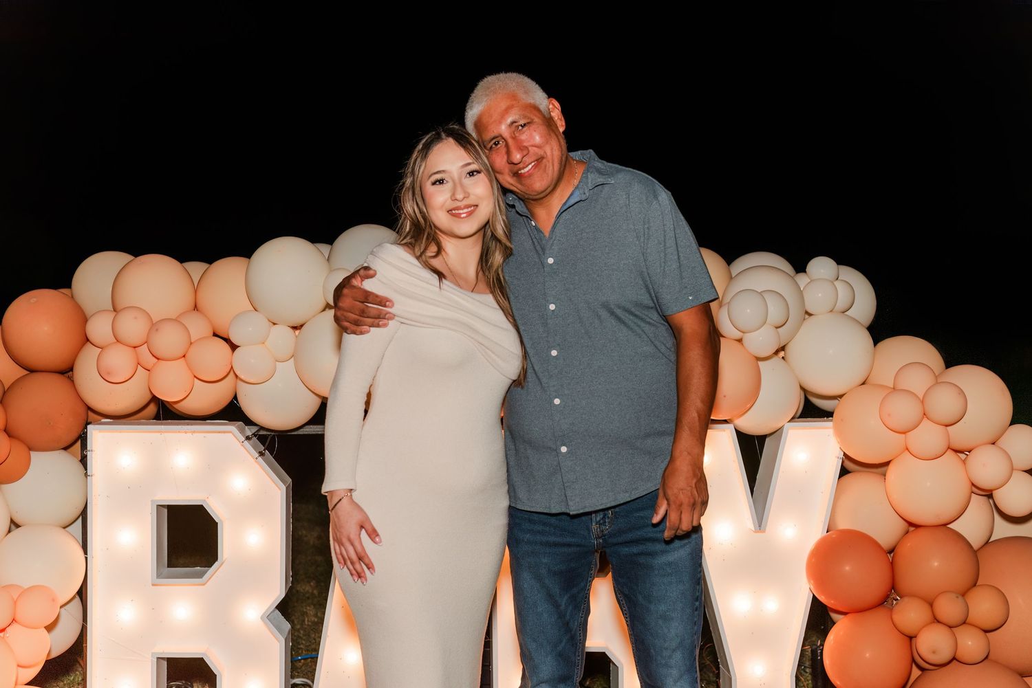A couple poses in front of a balloon arch and large letters at a party celebration.