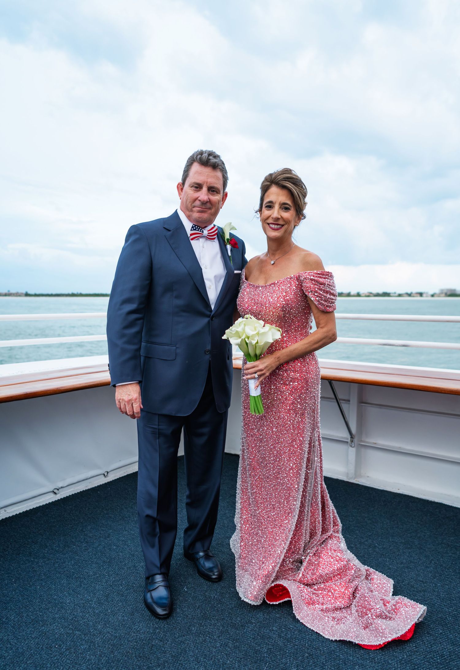Couple in formal attire posing on cruise ship deck, man in navy suit, woman in pink sequin gown holding white calla lily bouquet.