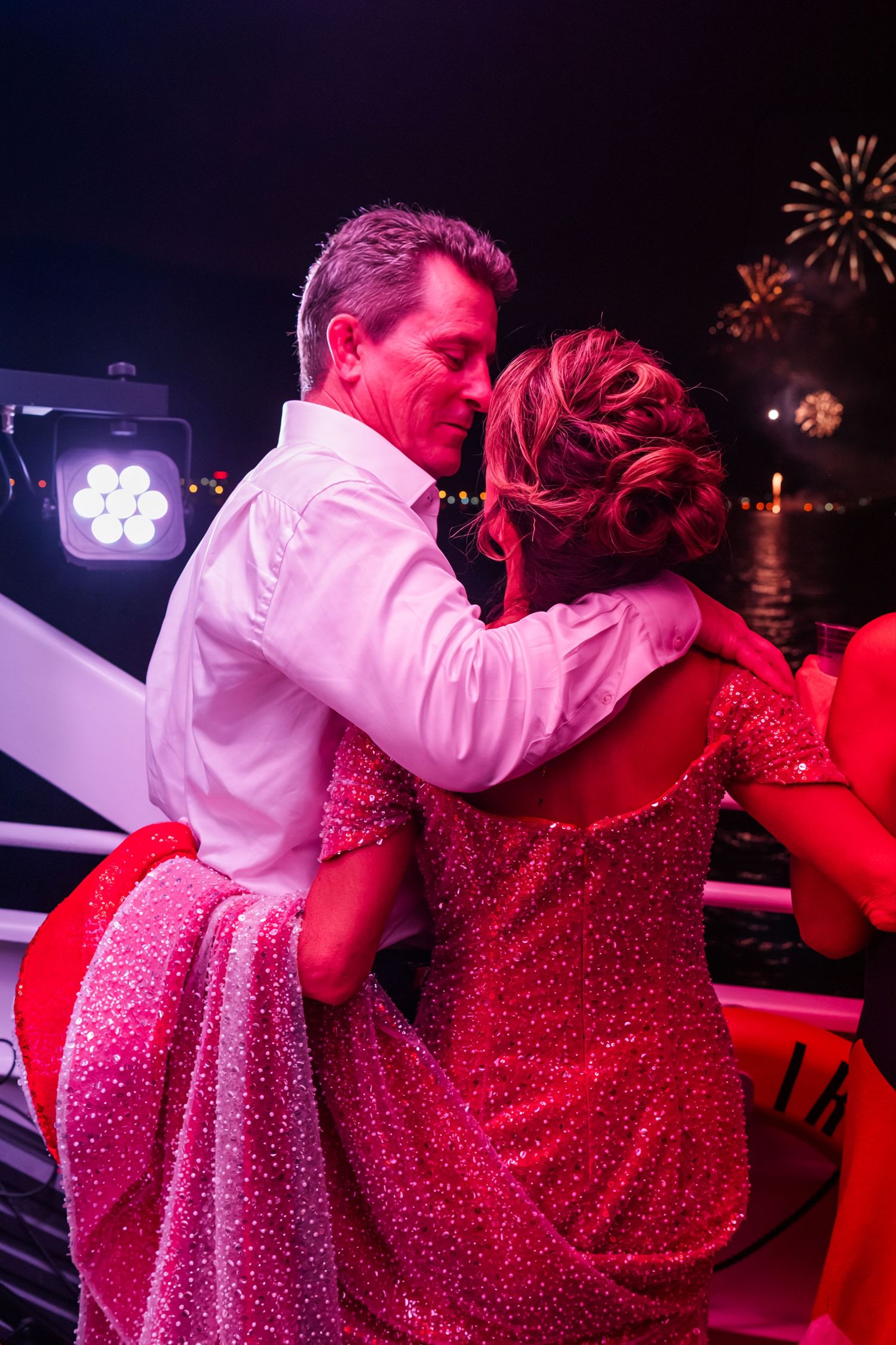 Couple embracing romantically on a boat deck with fireworks lighting the night sky, woman in red sequin gown.