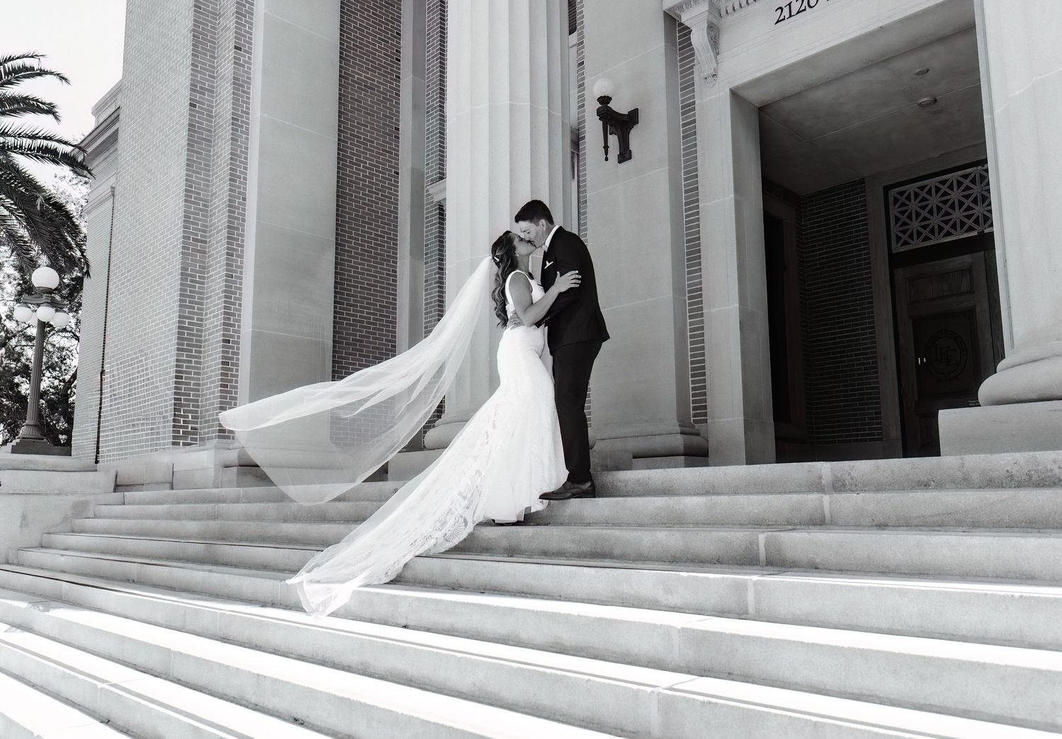 Black and white photo of a bride and groom sharing a kiss on church steps, veil flowing in the wind.