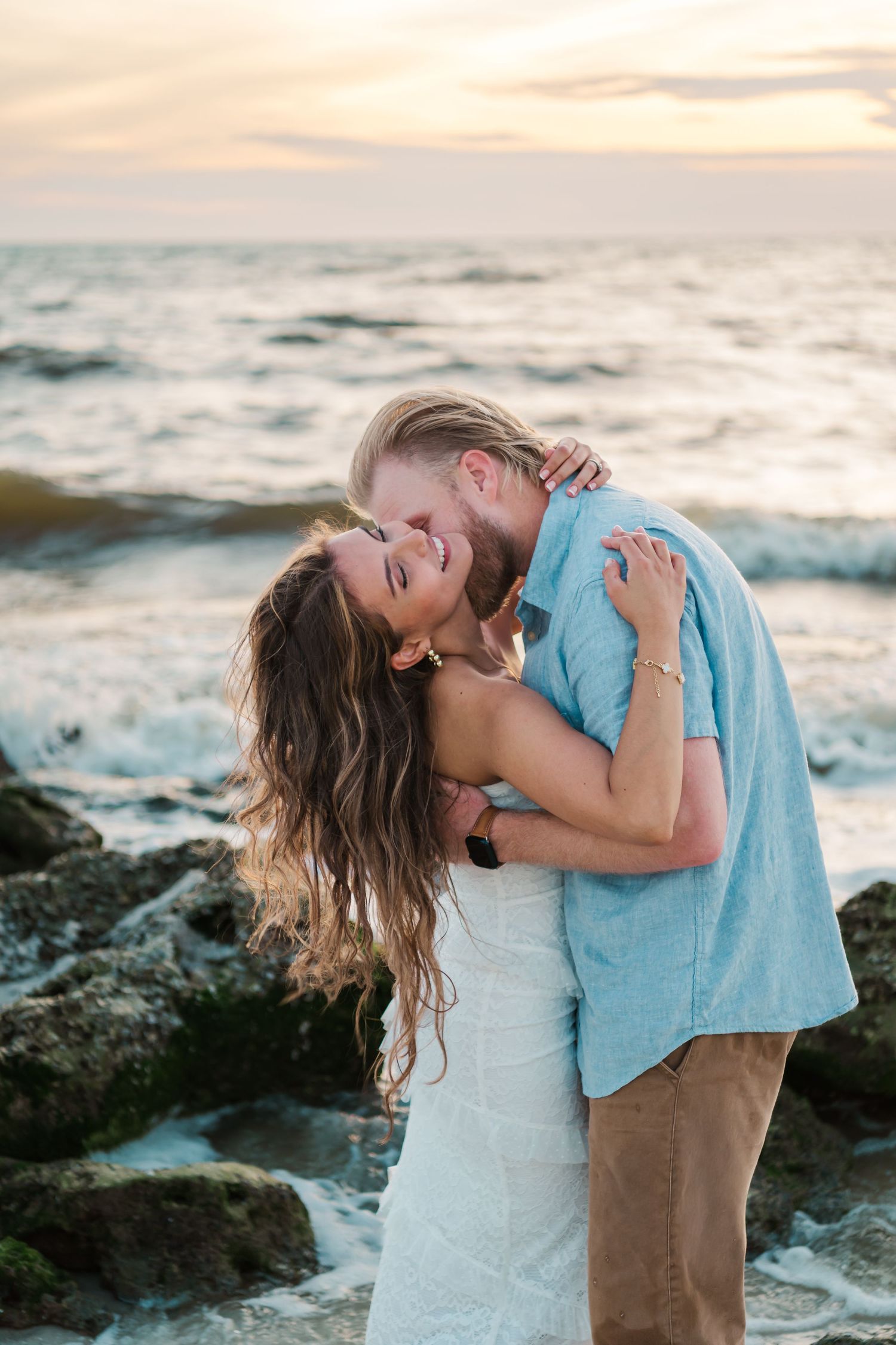 Couple embracing and laughing on rocky beach at sunset, woman in white dress, man in blue shirt.