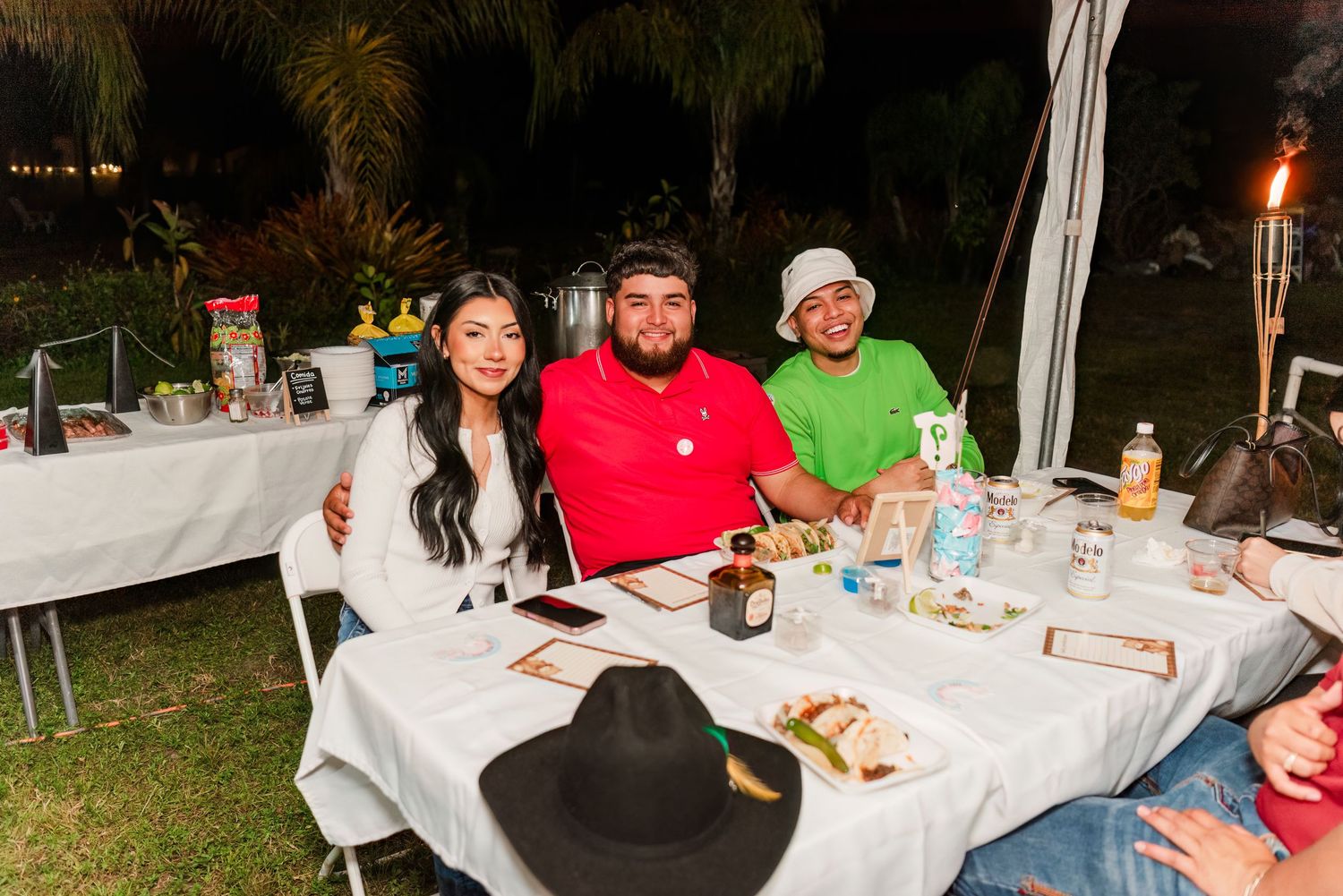 A group of people gathered around outdoor dining tables at night with festive lighting and table settings.