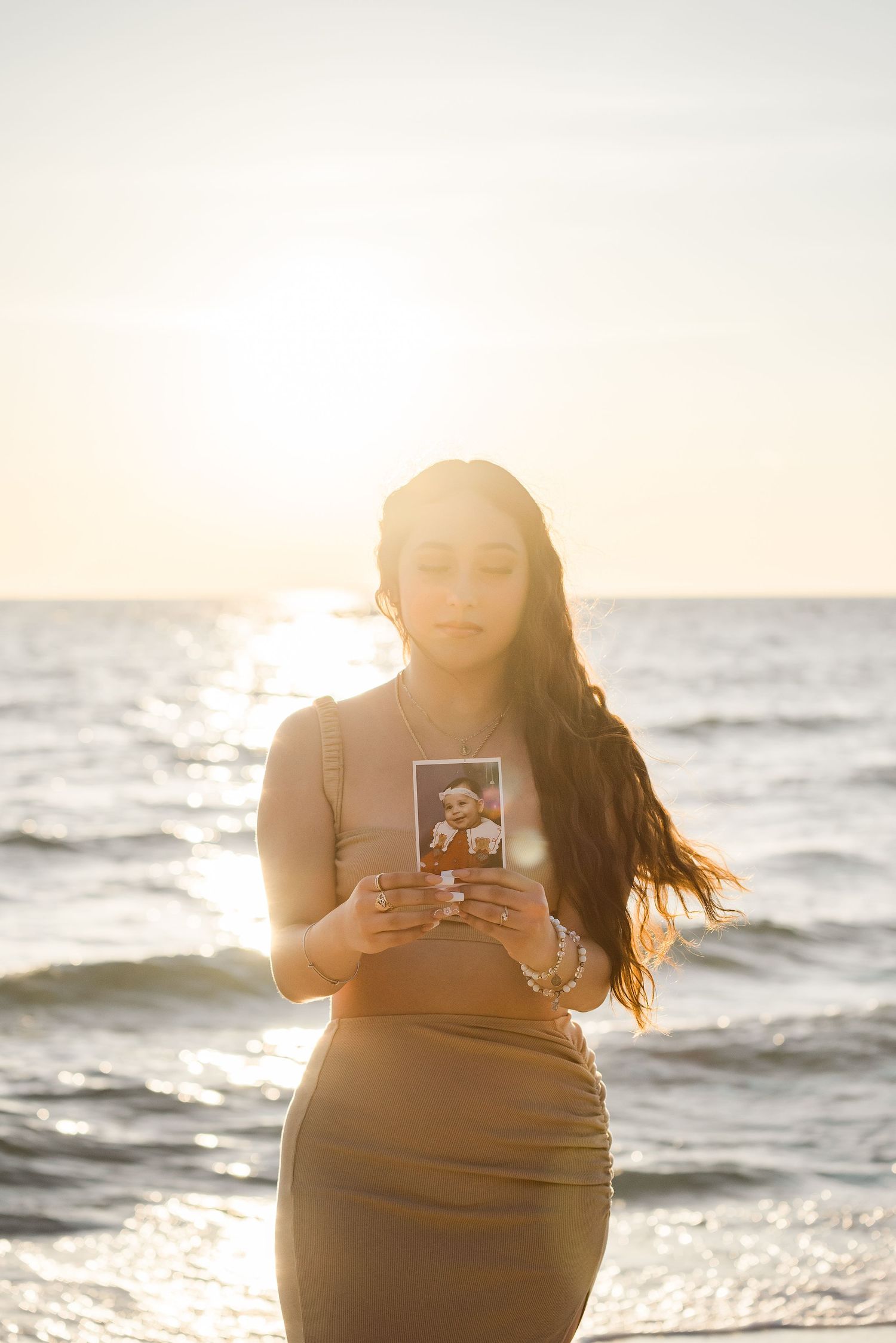 Person in brown dress holding a photograph while standing on beach at sunset with ocean waves in background.