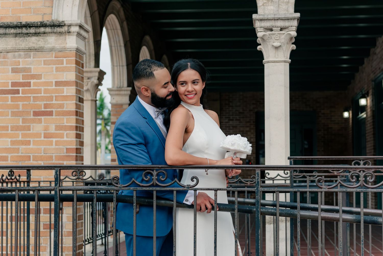Couple in formal attire embrace on a balcony with white columns and brick walls in an elegant architectural setting.