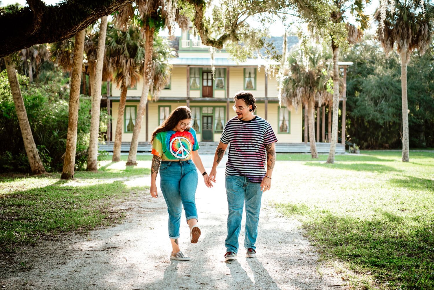 A couple walks hand in hand along a shaded path with Spanish moss and a historic house in the background.
