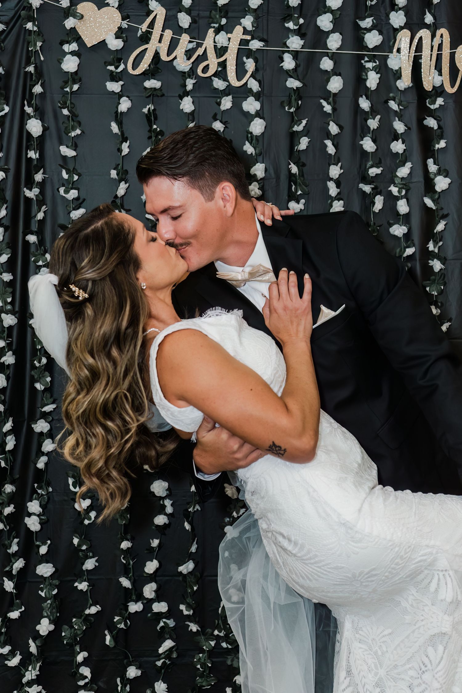 Bride and groom share a romantic dip kiss in front of a black floral backdrop with a Just Married sign.