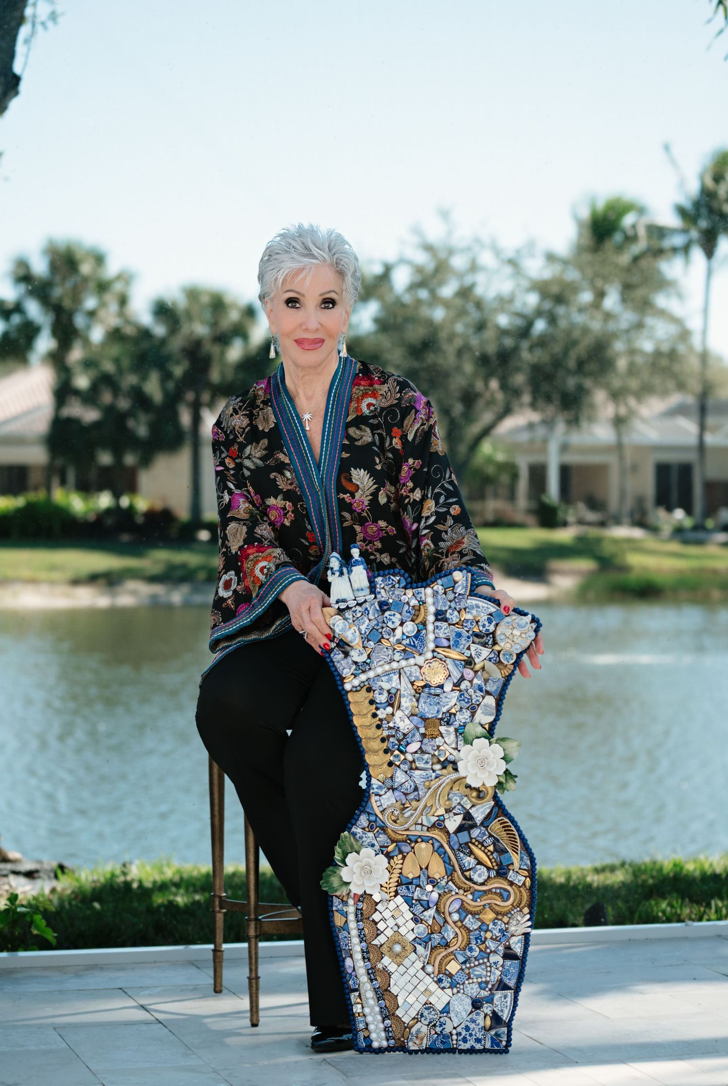 Stylish older woman in floral kimono poses outdoors beside a blue and white mosaic guitar sculpture near a lake.