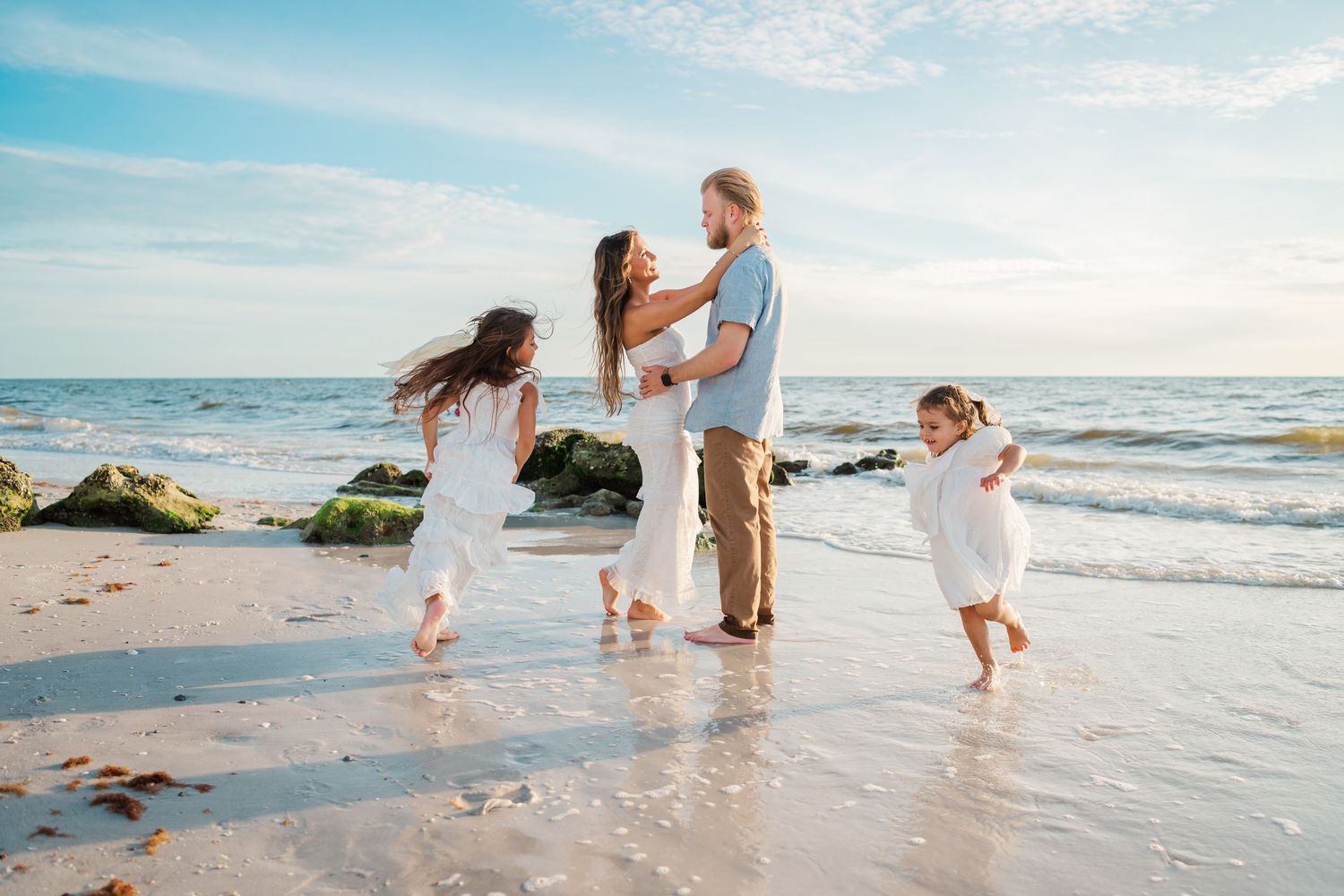 Family in white outfits playing on a sunny beach with waves, rocks, and a blue sky backdrop.