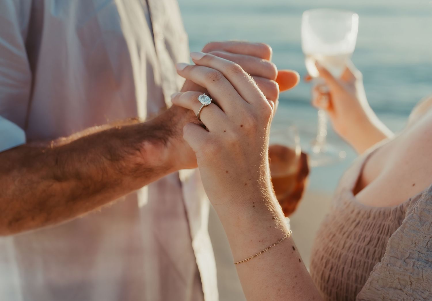 Two hands holding champagne glasses together in celebration with a sparkly ring visible against a soft blue background.