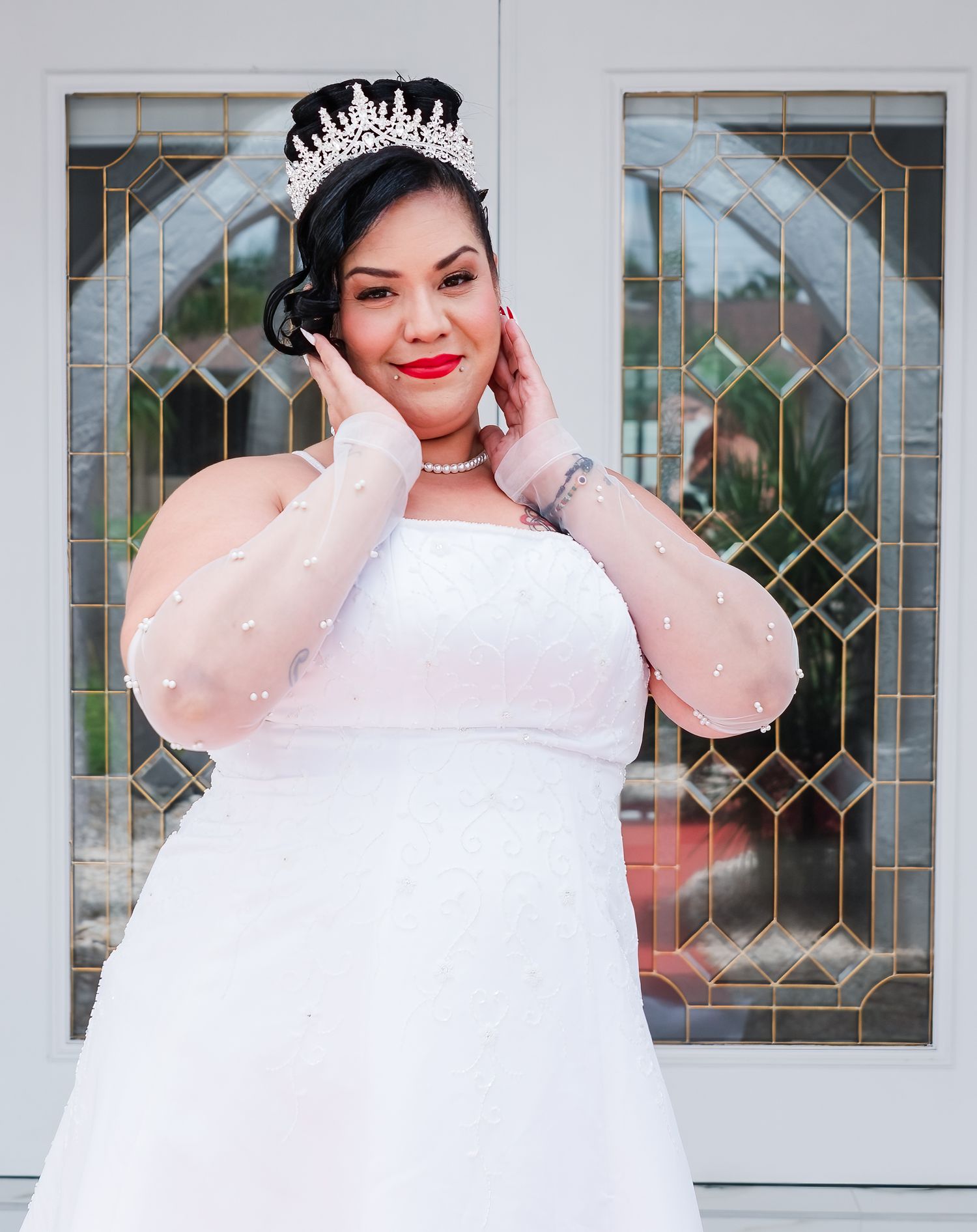 A smiling bride in a white wedding gown and tiara poses elegantly against leaded glass windows.