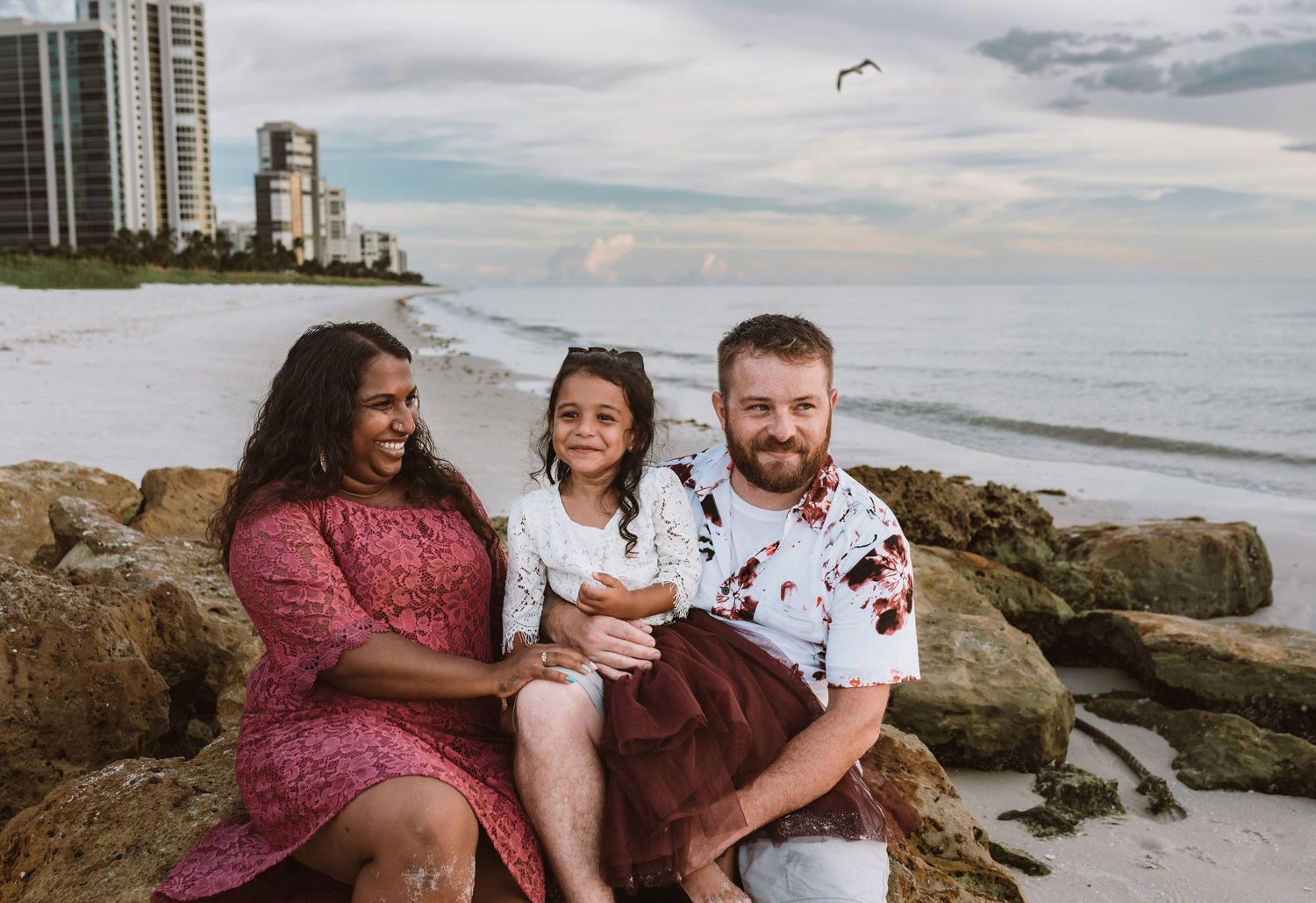 A family sits together on coastal rocks at the beach with high-rise buildings and cloudy skies in the background.