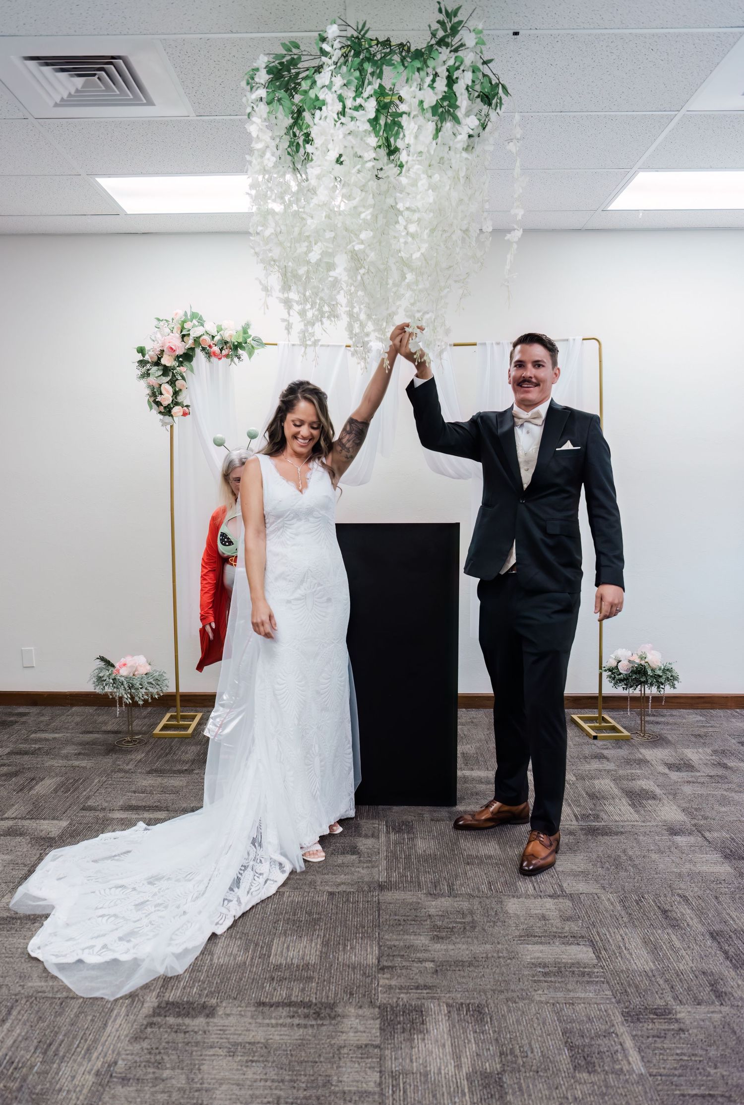 Joyful newlyweds raise hands at wedding ceremony beneath floral chandelier with gold arch backdrop.