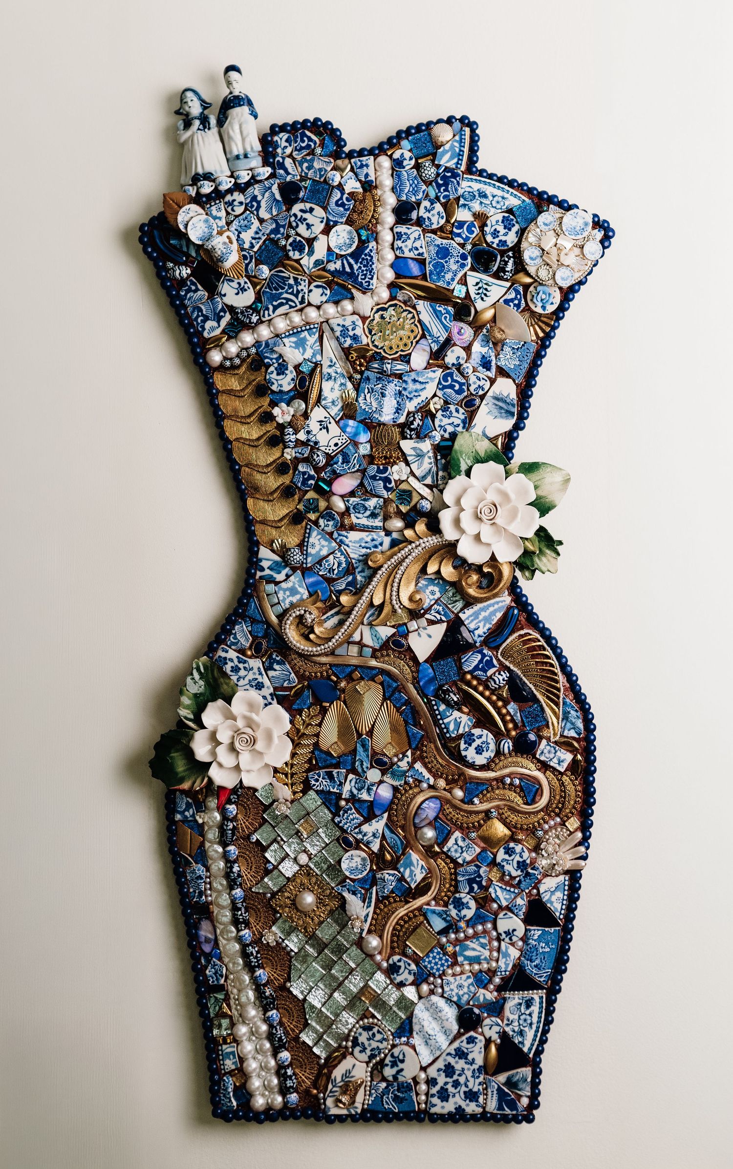 Blue and white mosaic dress sculpture adorned with pearls, ceramic flowers, and figurines in Delft-inspired pattern.