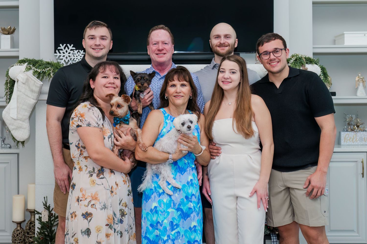 Family of seven posing together indoors for a holiday photo, holding small dogs near a festive fireplace mantel.