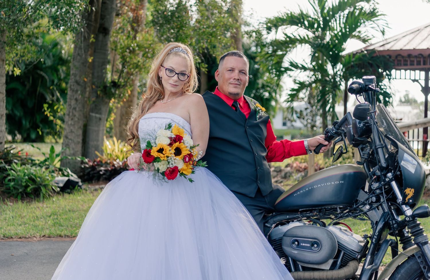 Newlyweds pose next to black motorcycle in garden setting with bride in white ballgown holding colorful bouquet.