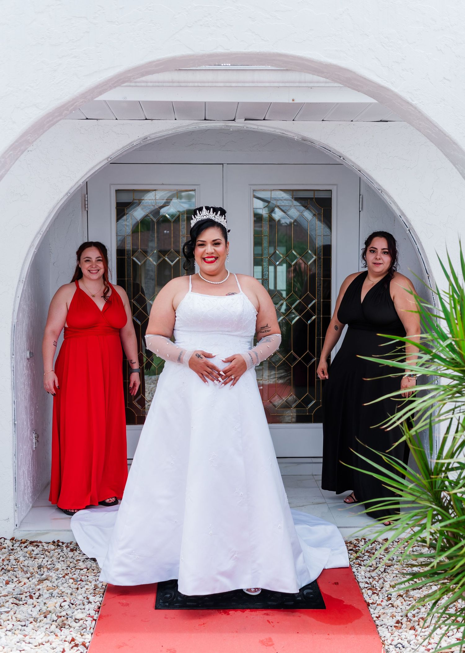 Three figures stand under an archway on a red carpet, with a person in a white gown center flanked by two in formal dresses.