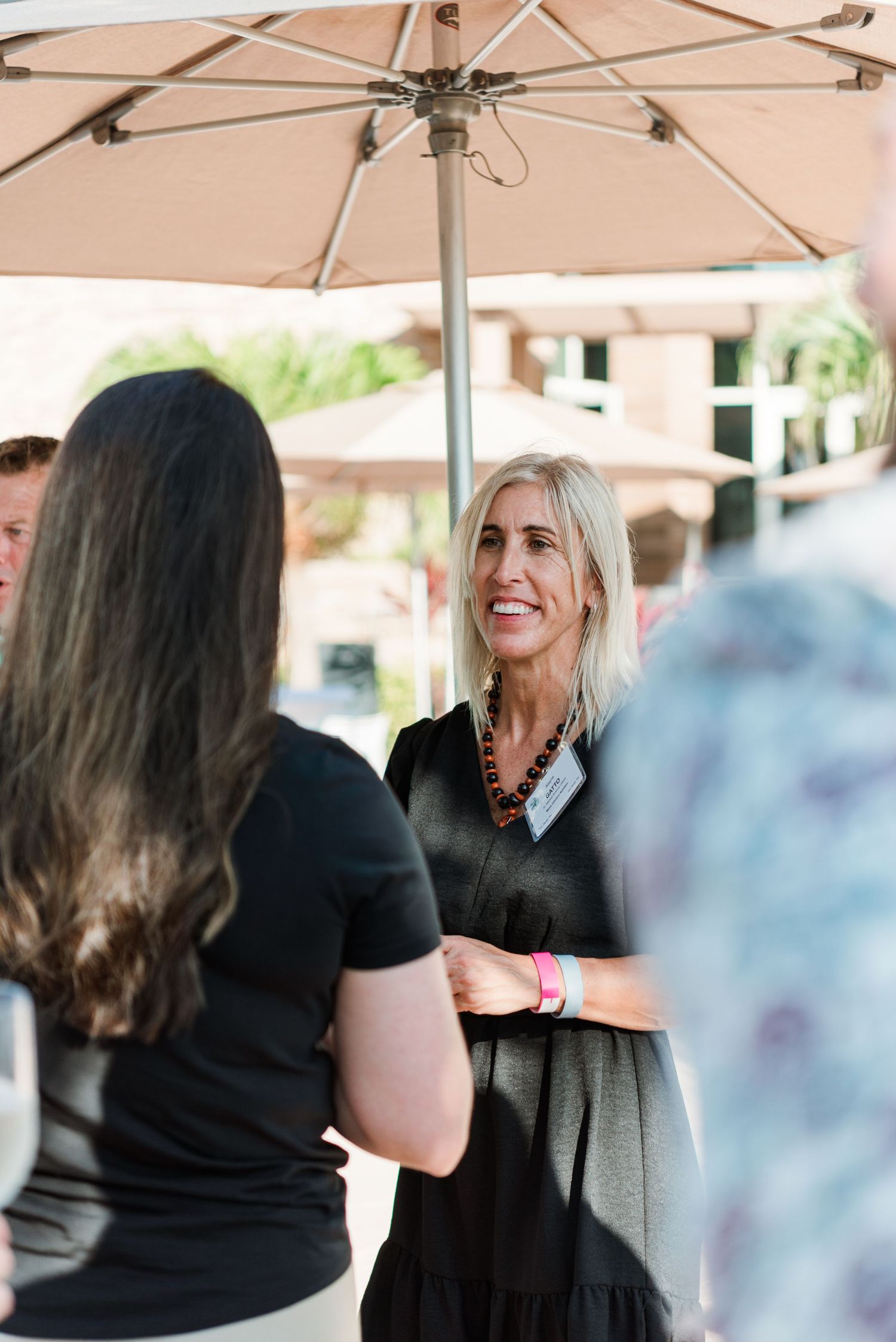 People having an animated conversation under a beige patio umbrella at an outdoor gathering.