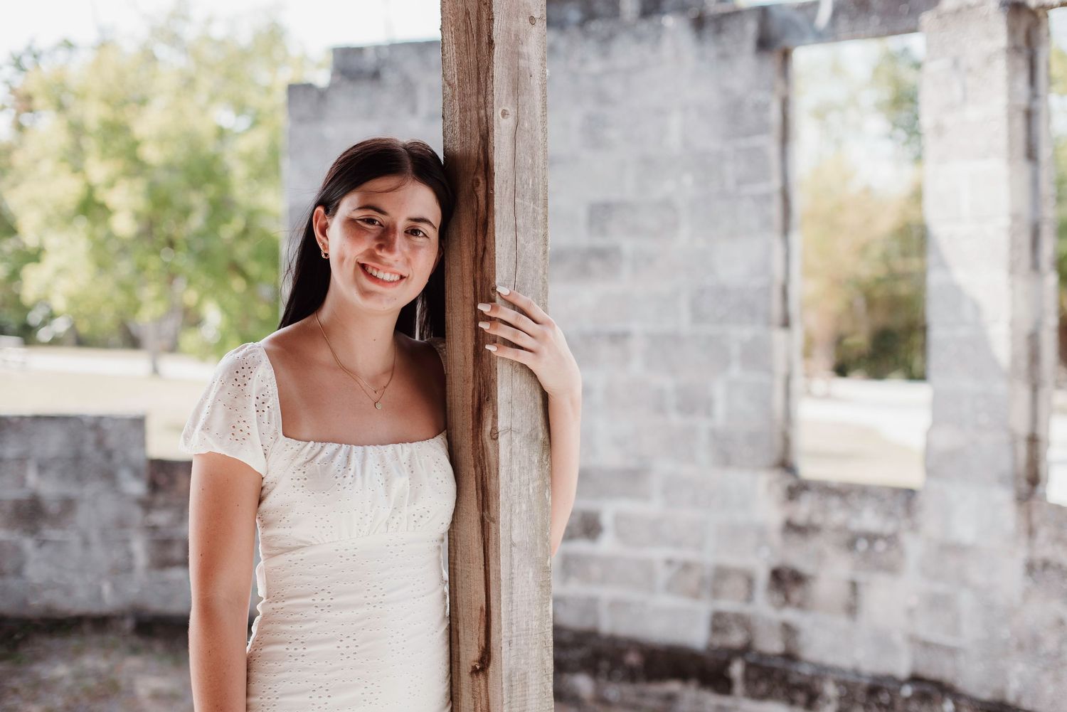 Woman in cream dress poses beside wooden post in front of rustic stone wall.