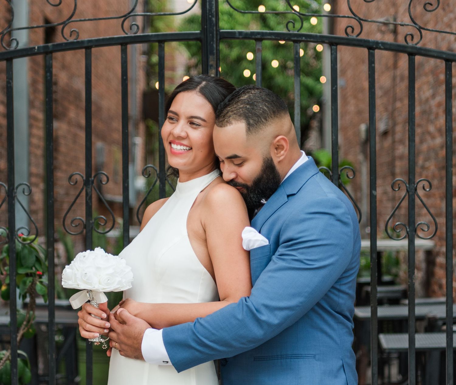 A romantic wedding couple embraces in front of an ornate iron gate with a lovely garden backdrop in warm sunlight.
