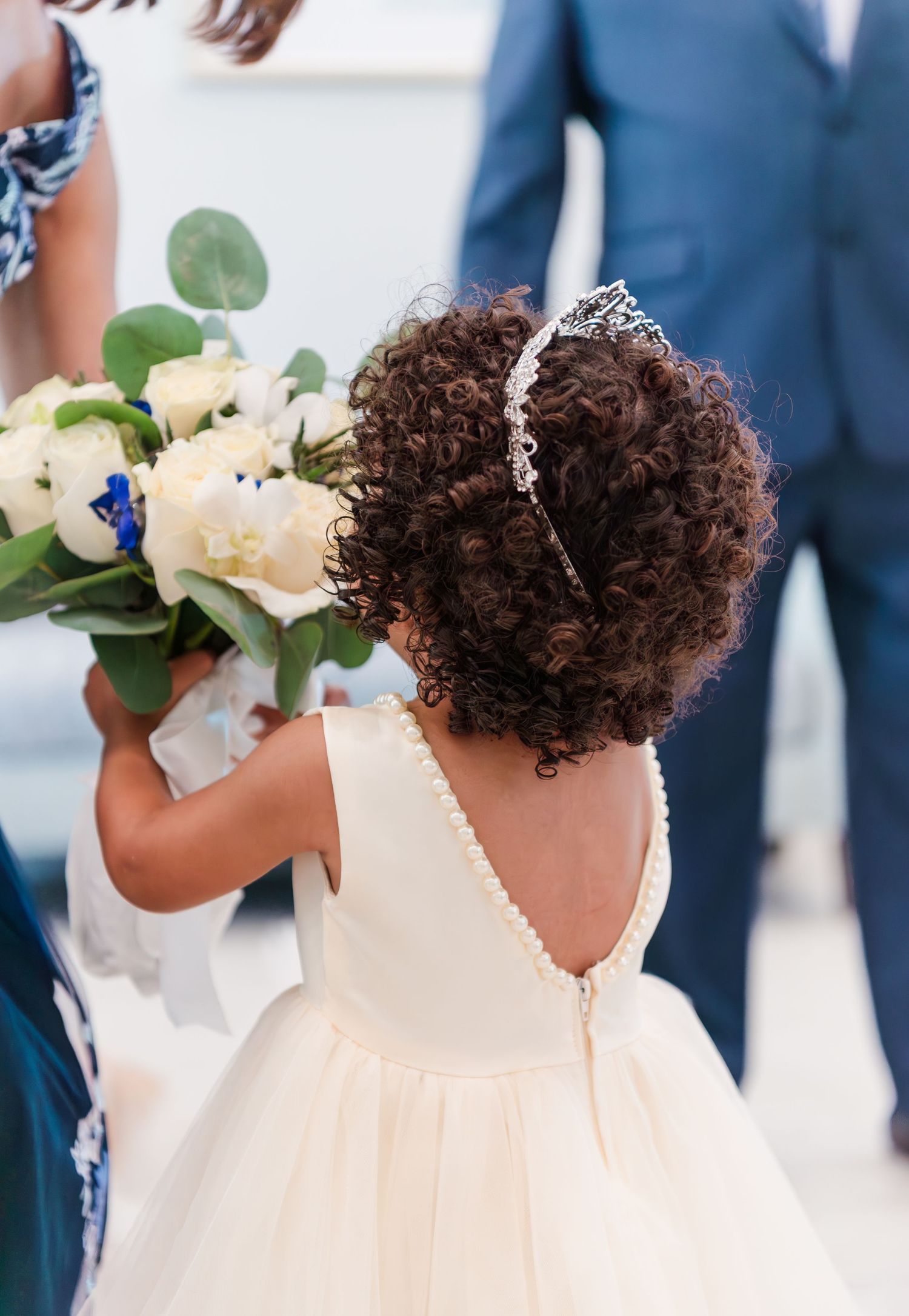 A young flower girl in a white dress with natural curly hair holds a bouquet of white and blue flowers at a wedding.