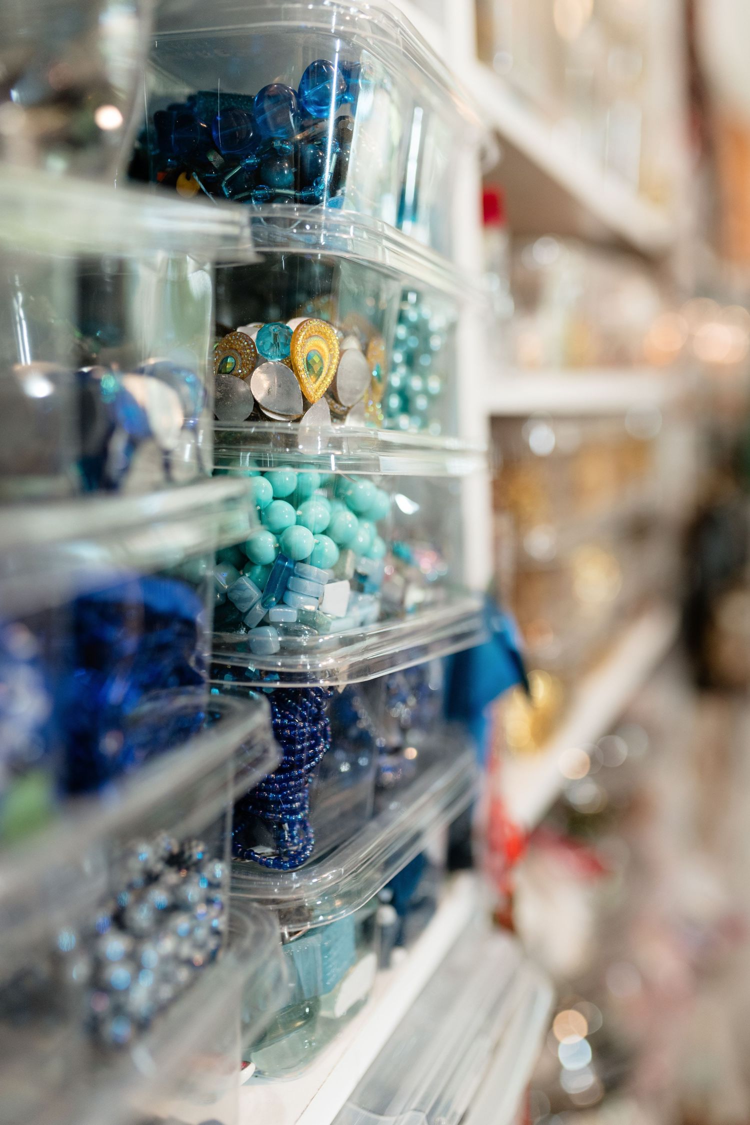 Clear plastic bins on white shelves display colorful beads in blue, teal, and turquoise shades in a craft supply store.