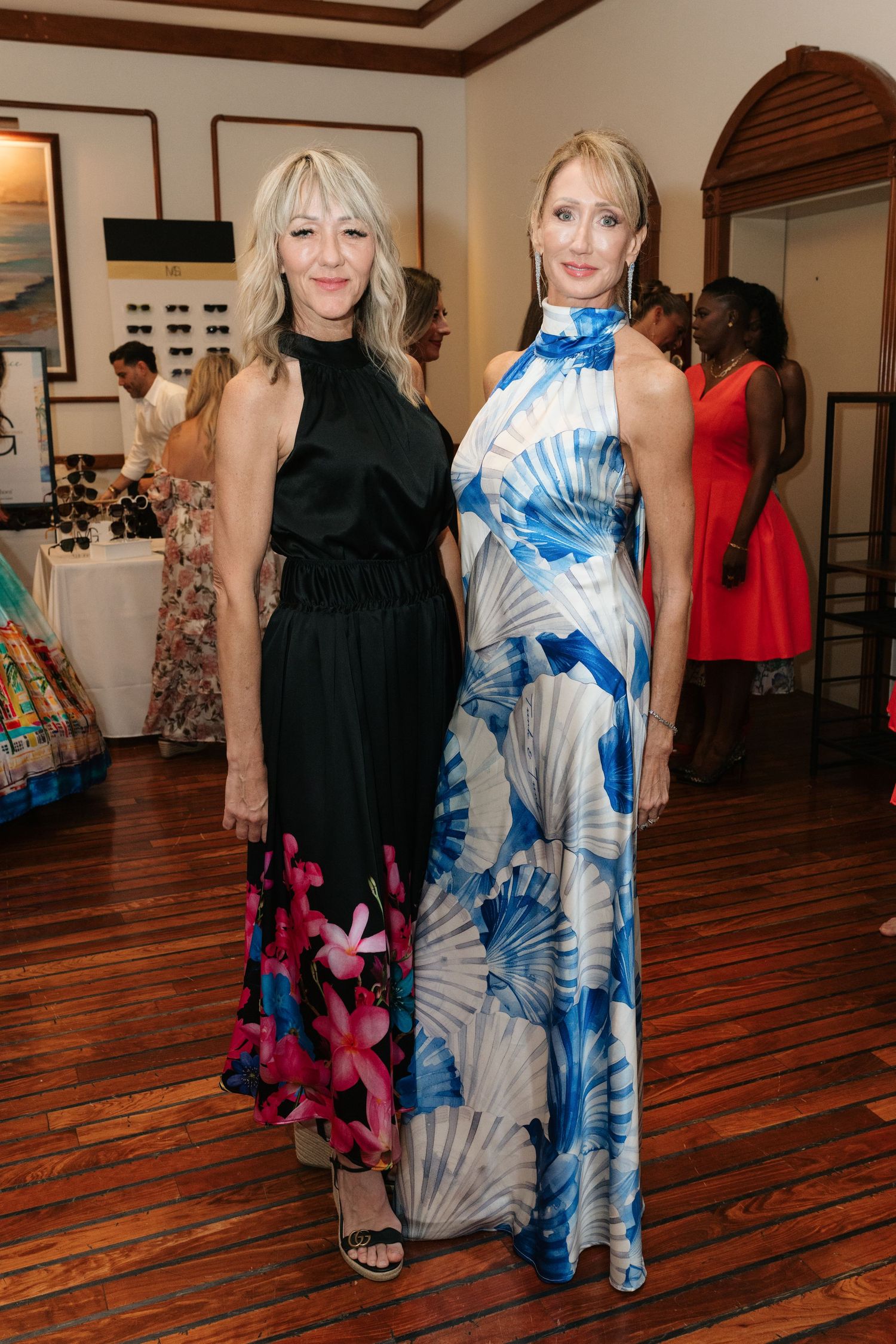 Women in elegant dresses, including a black gown and blue-and-white patterned dress, standing on hardwood floors at an event.