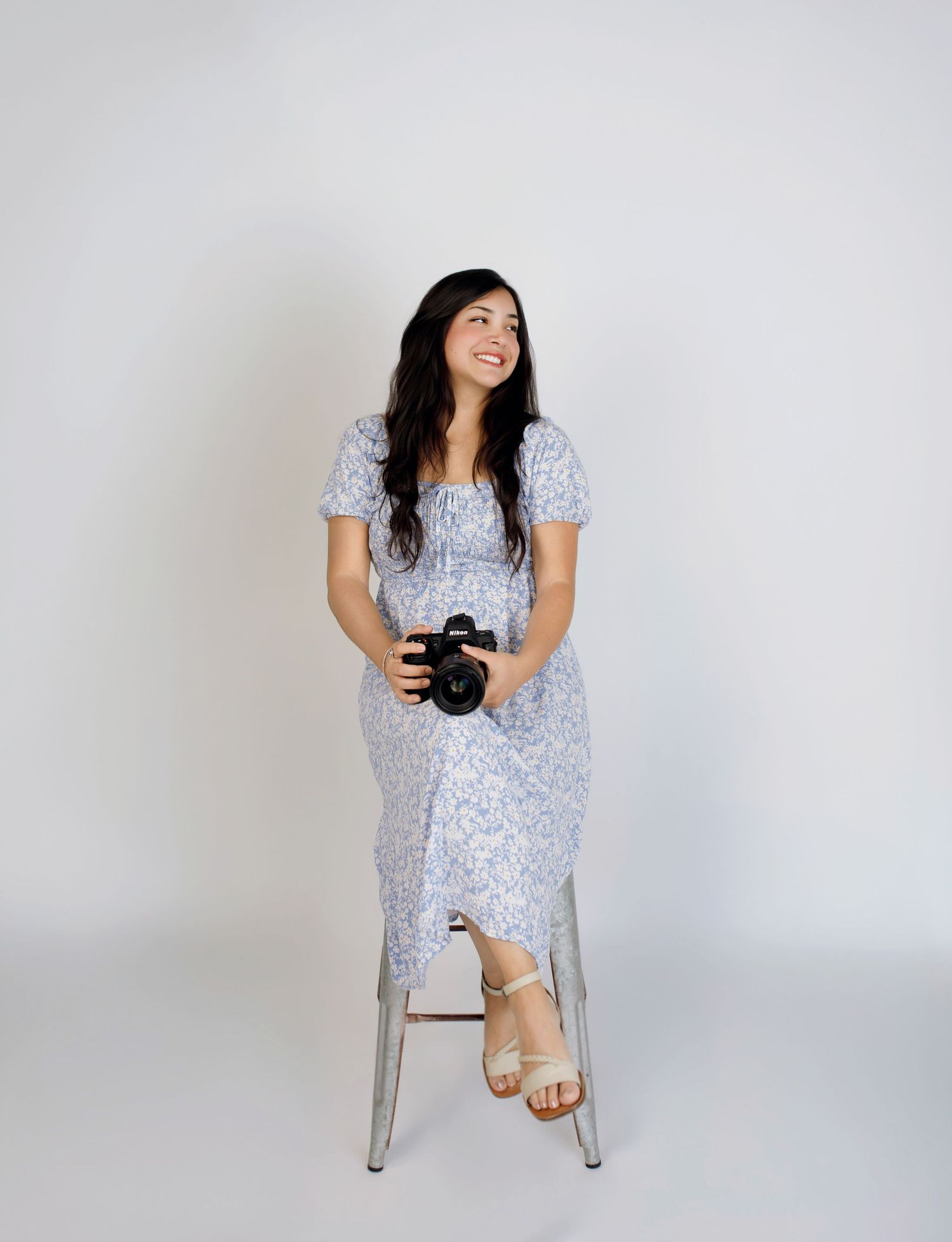 Woman in floral blue dress sitting on stool holding camera, smiling upward against white background.