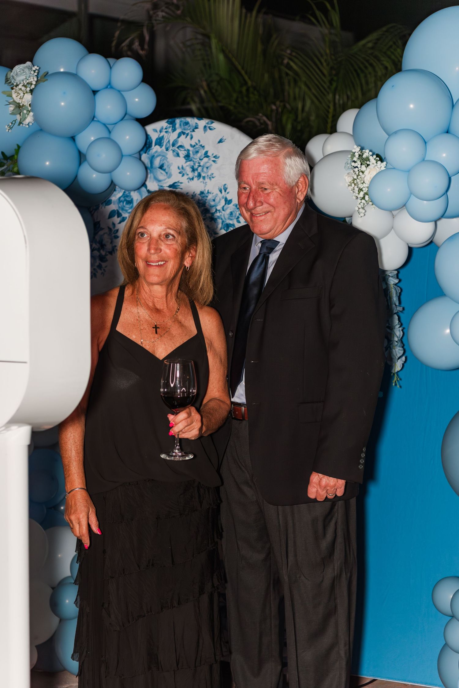 A couple poses in formal attire against a backdrop of blue balloon decorations at an elegant event.