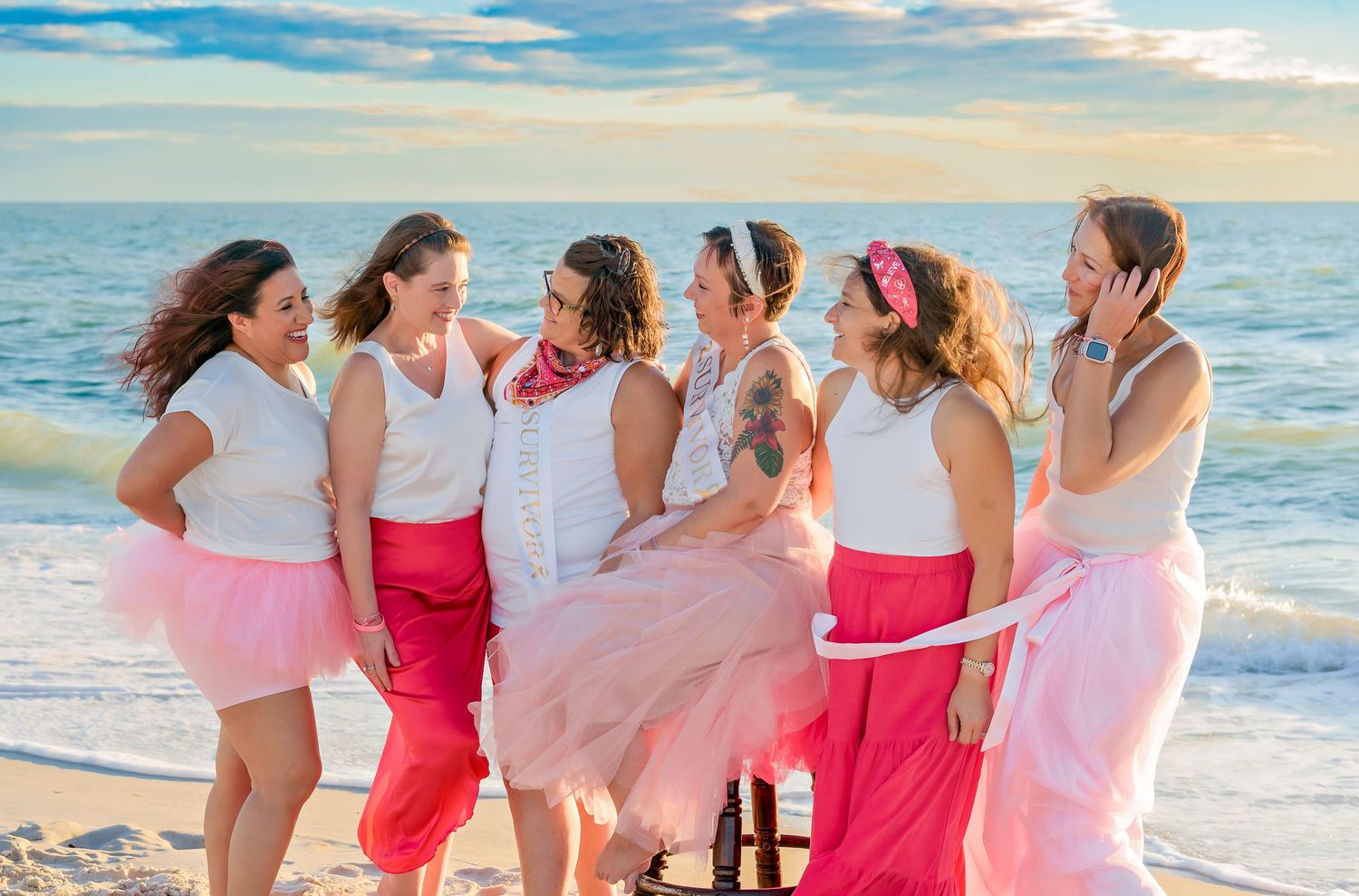 Group wearing pink and coral dresses walking together on a sunny beach at sunset.