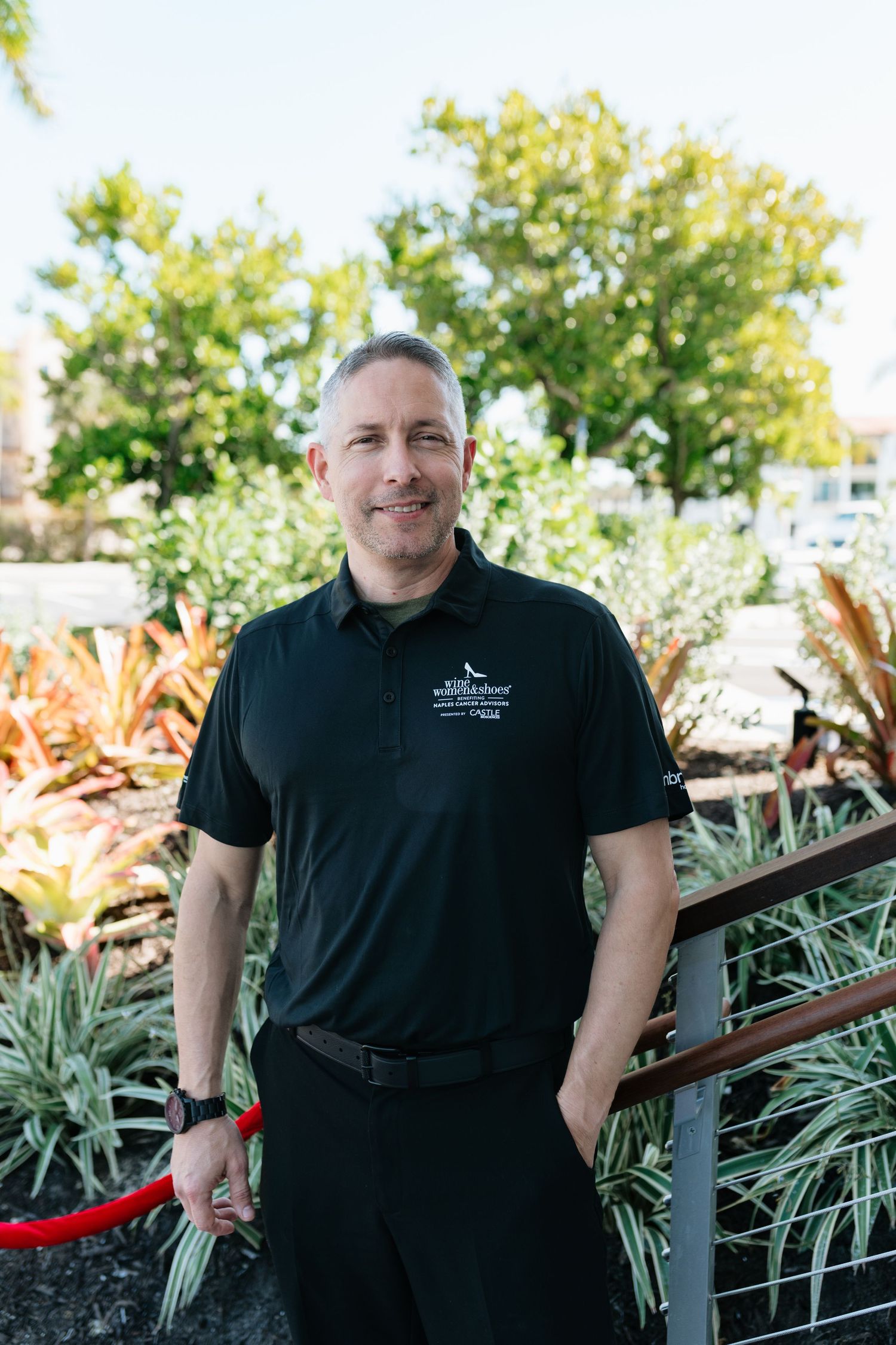 Person wearing black attire poses in an outdoor garden setting with lush tropical plants and greenery.
