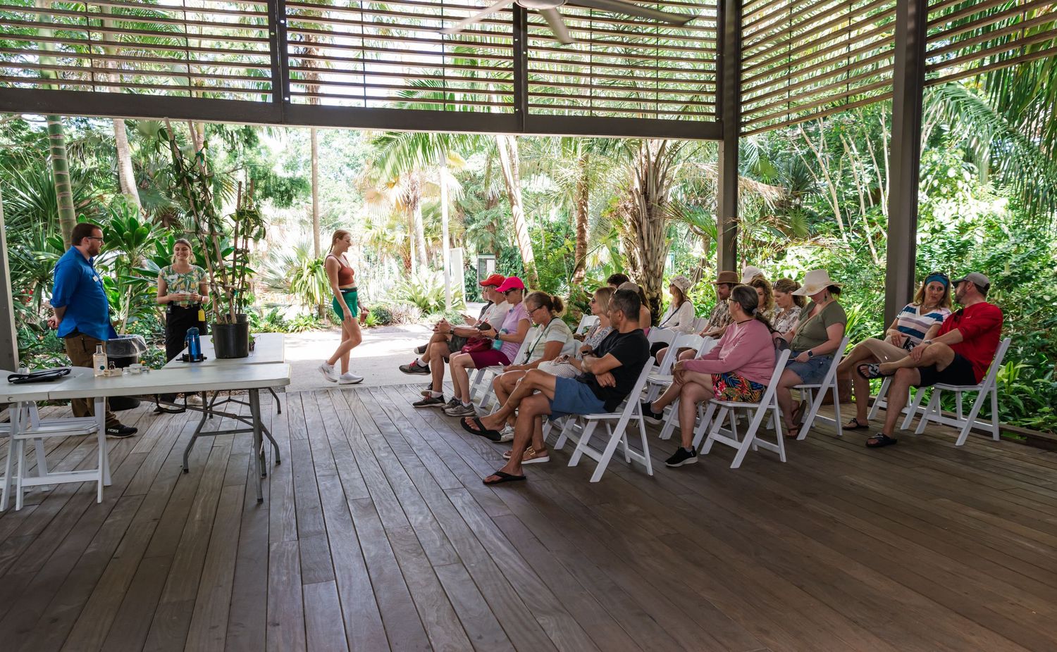 Group of tourists attending an outdoor presentation in a tropical open-air pavilion surrounded by lush greenery.