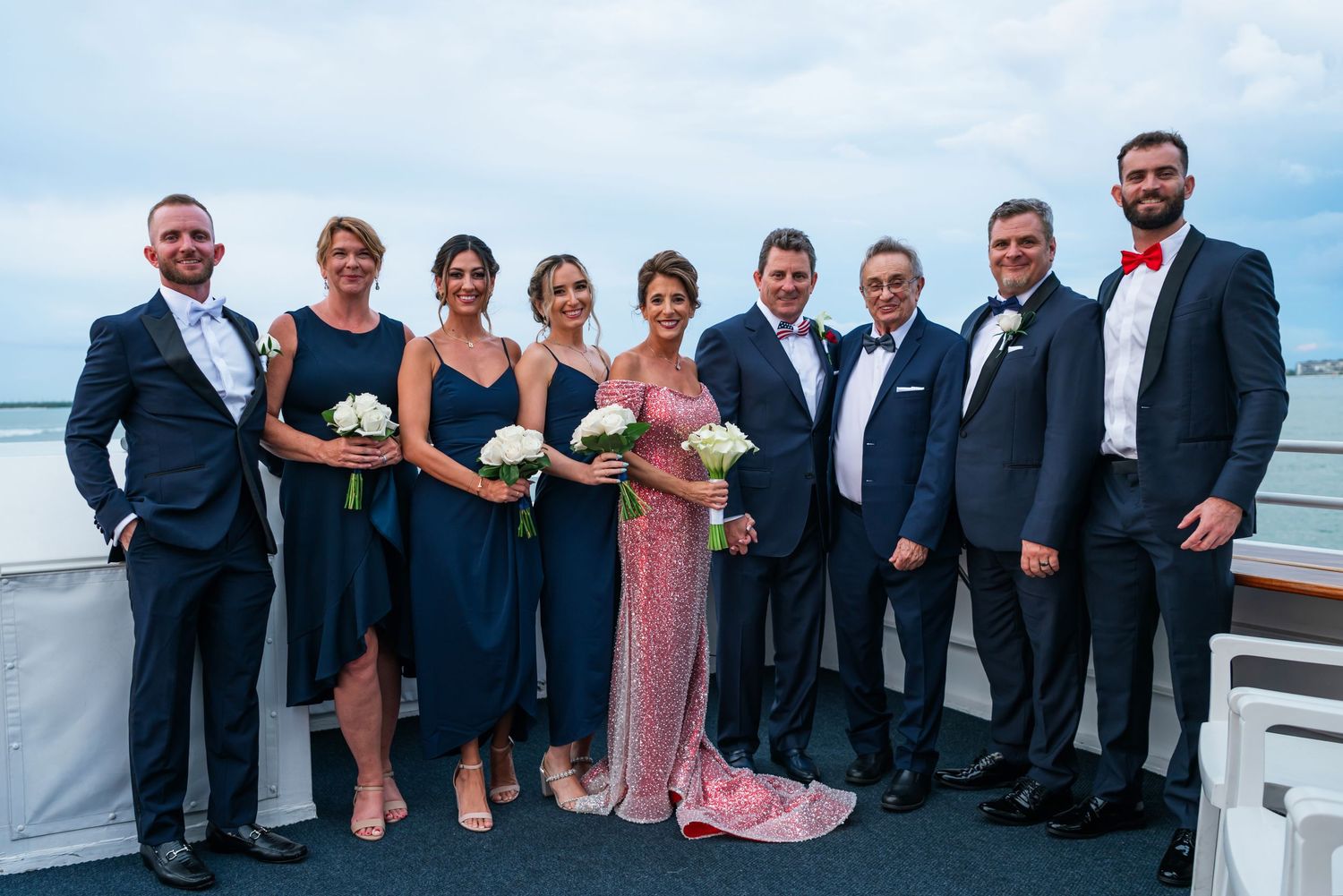 Wedding party posing on a boat deck, wearing navy suits and dresses, with bride in pink gown holding white bouquet.