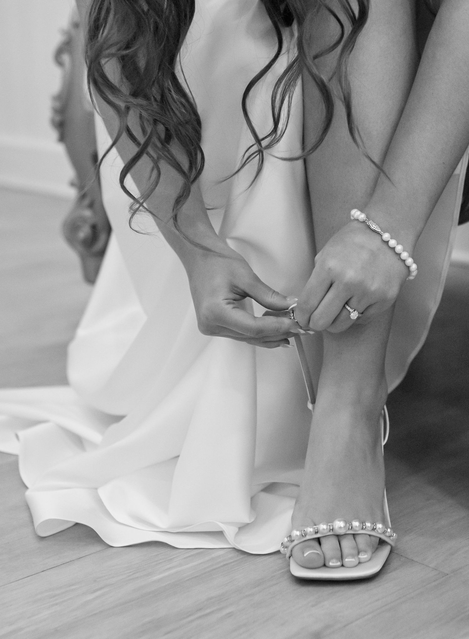 Bride fastening pearl-adorned heels in a white wedding gown, wearing a pearl bracelet and ring, black and white photo.
