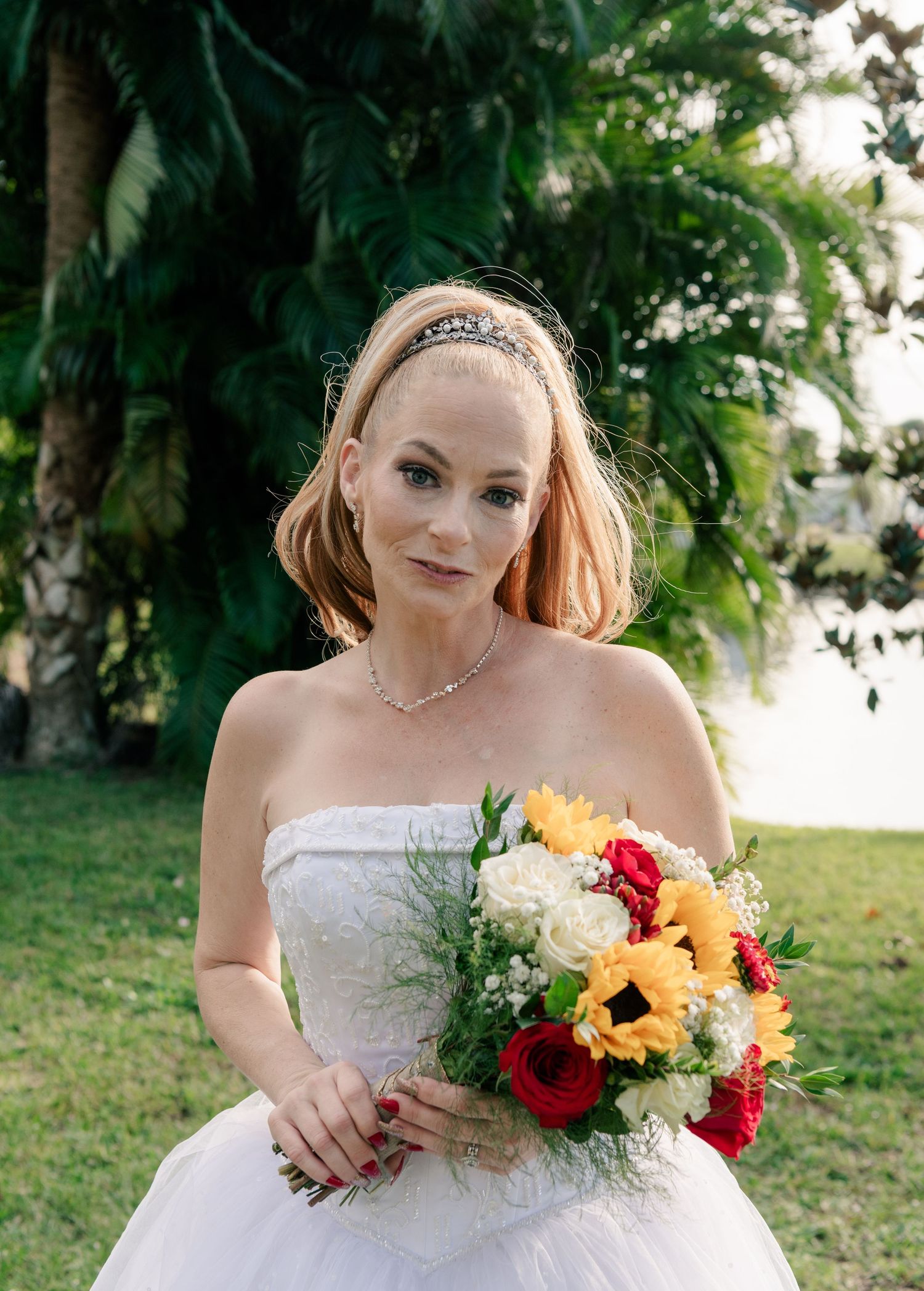 A bride in a strapless white gown holds a vibrant bouquet of sunflowers and red roses against a tropical garden backdrop.