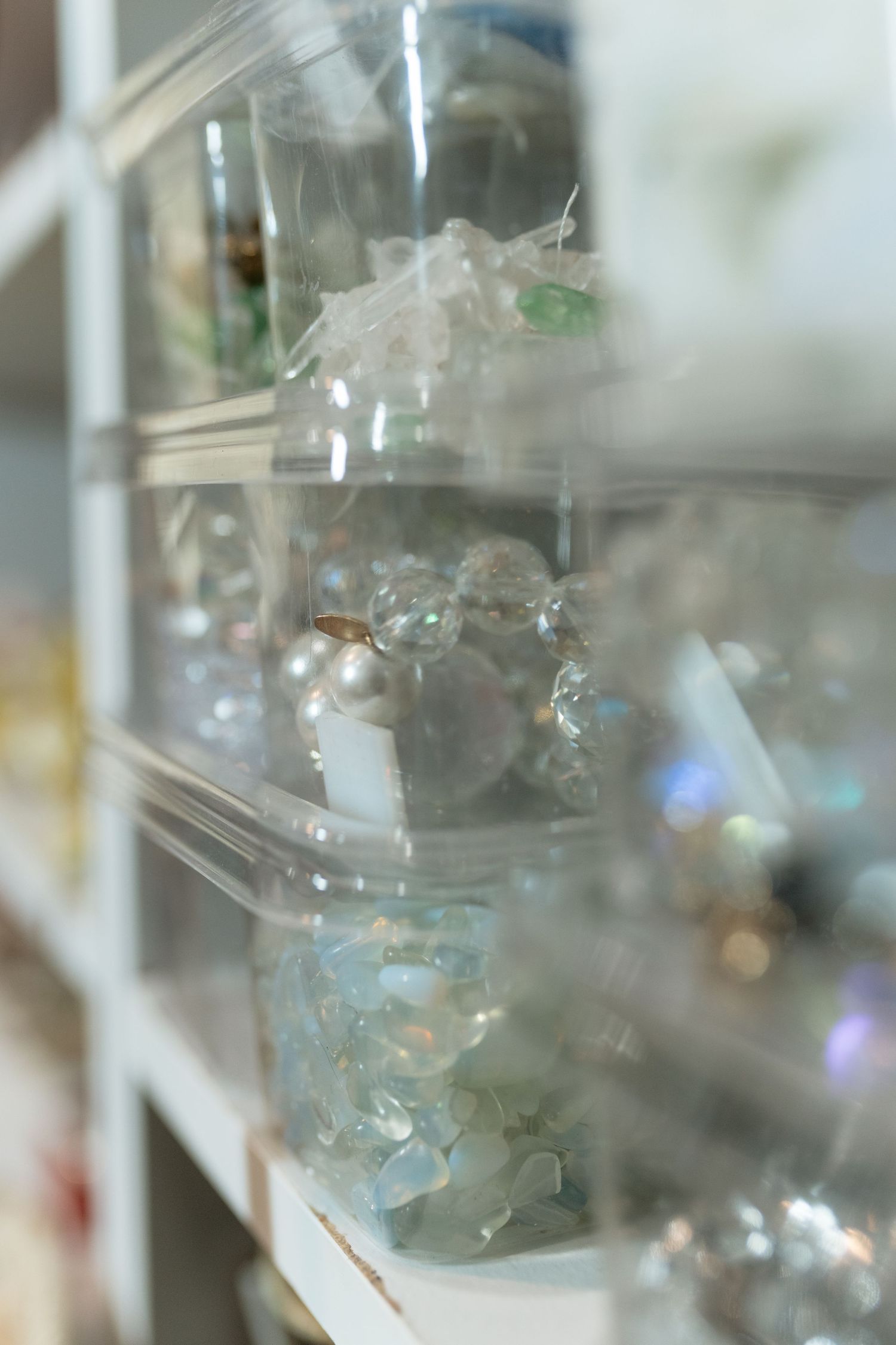 Clear plastic bins on white shelves display assorted crystals, pearls, and gemstones in a jewelry or craft store.