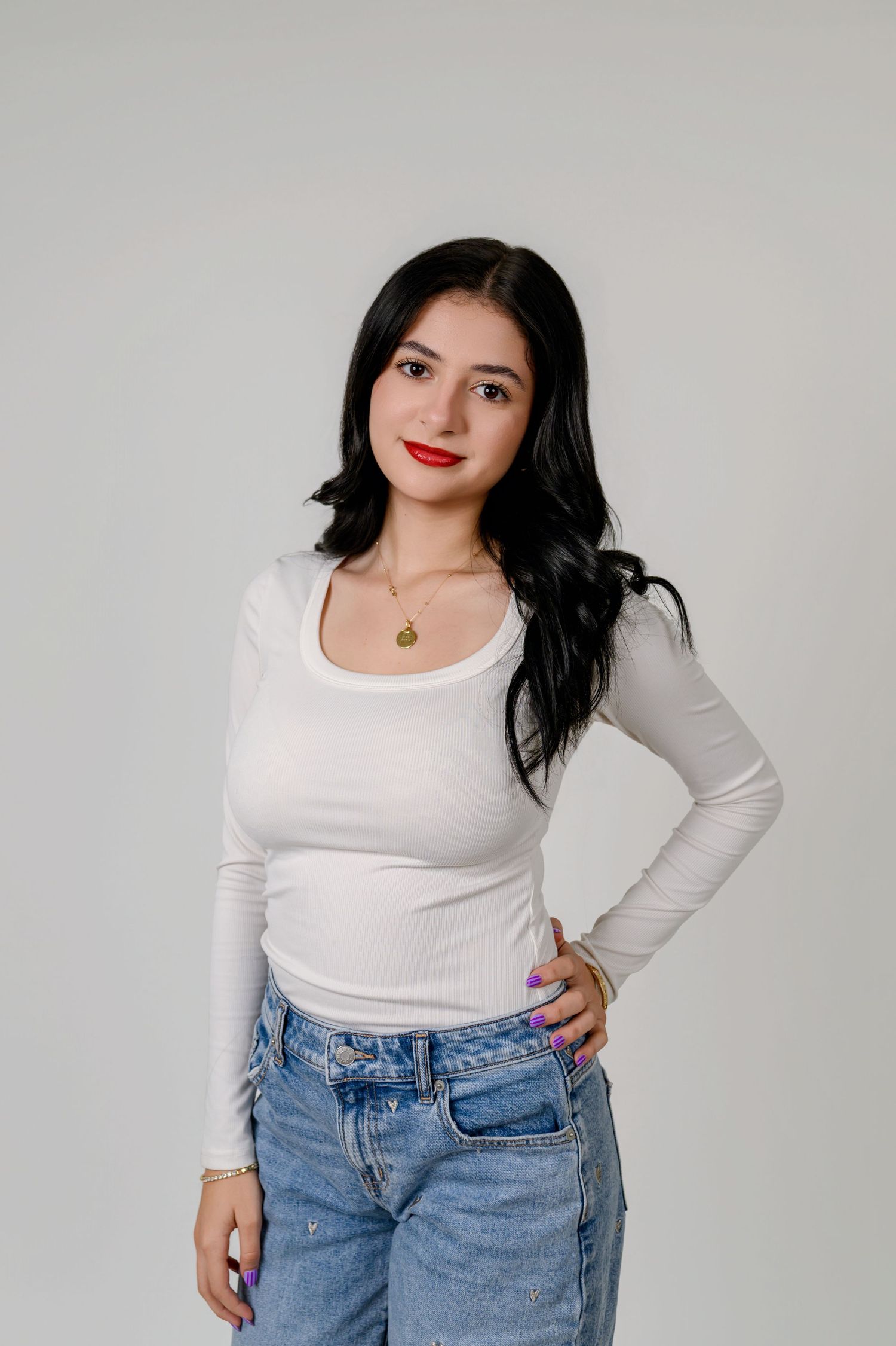 Young woman with dark hair in white long-sleeve top and jeans poses confidently against a light gray background.