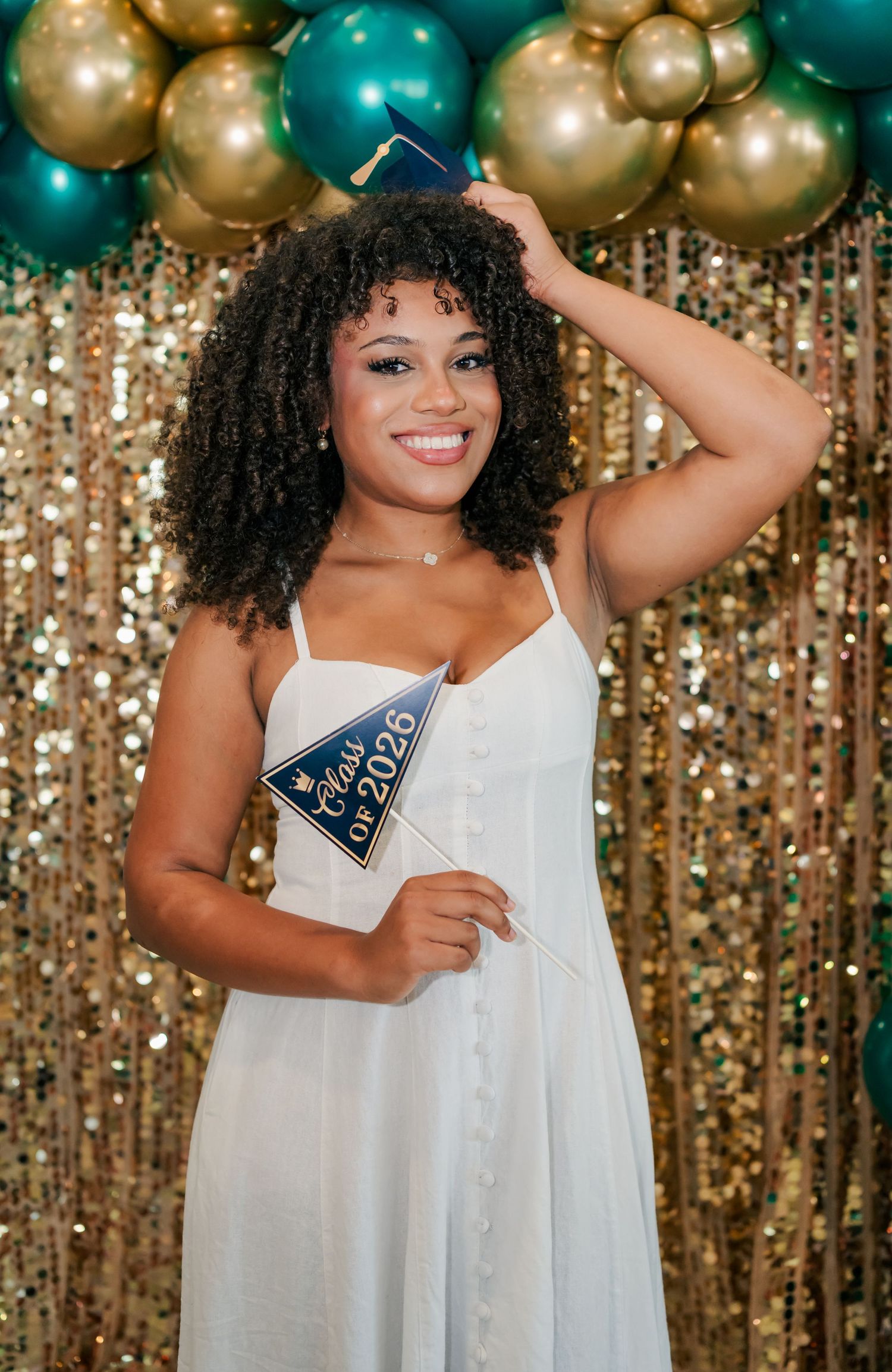 Smiling graduate in white dress holds Class of 2025 pennant with mini cap, posing before gold sequin backdrop with teal and gold balloons.