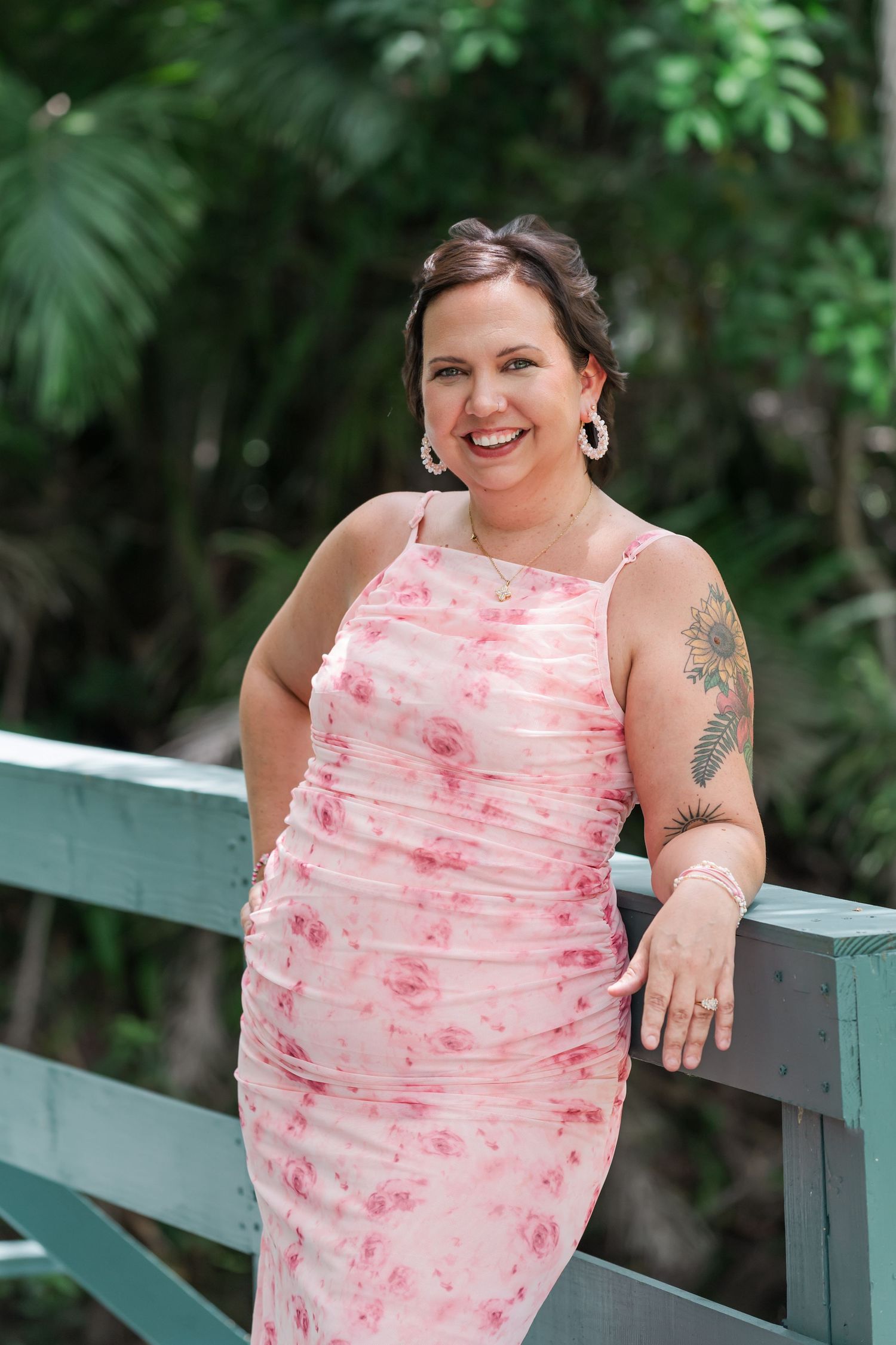 Woman in pink floral dress smiling on bridge surrounded by lush greenery with tattoo visible on arm.