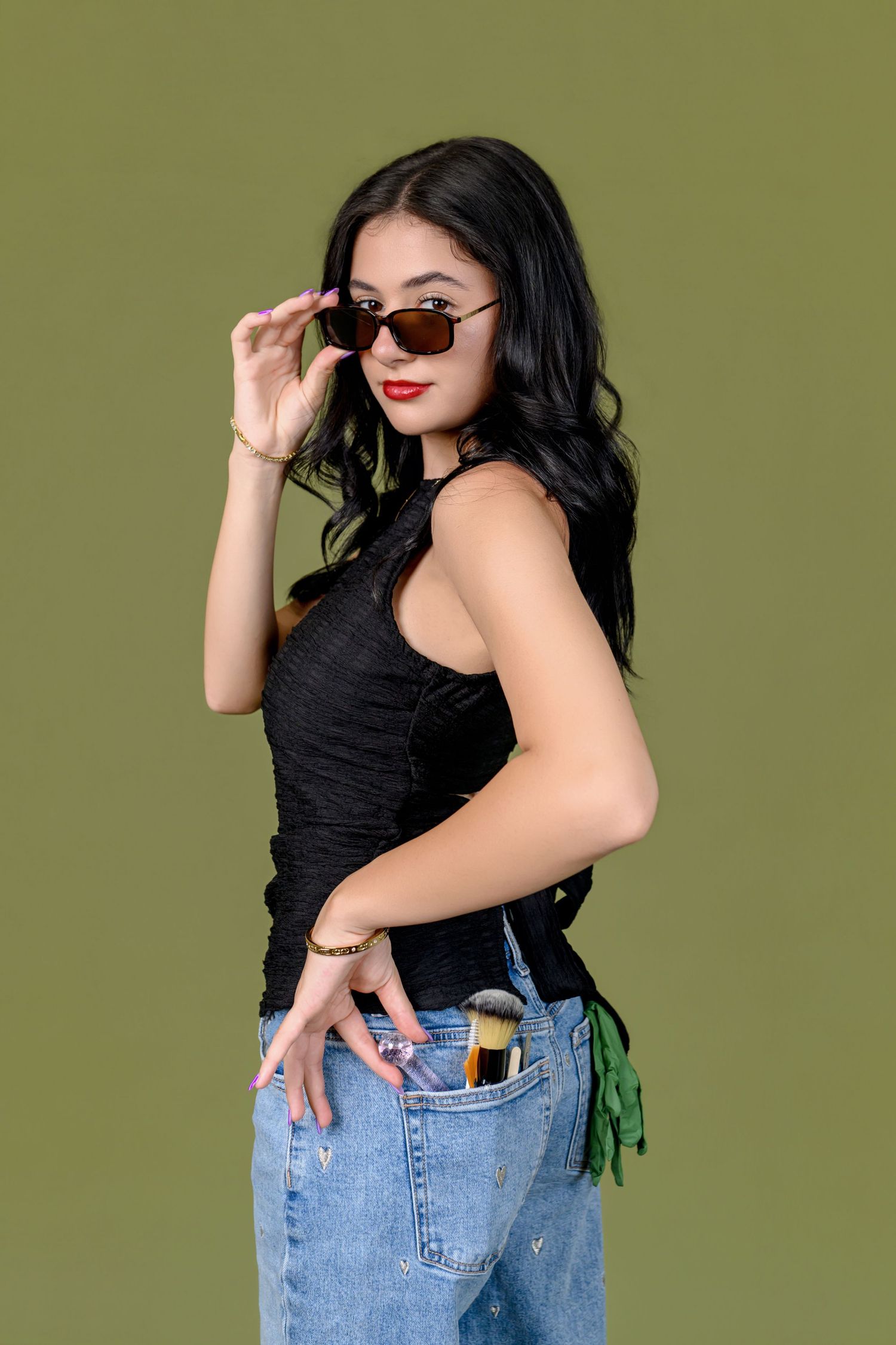 Young woman in black tank top and jeans, wearing brown sunglasses, posing against an olive green background.