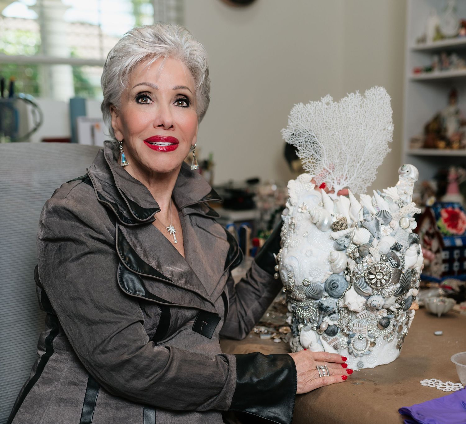 Elegant older woman with silver hair displays ornate white seashell and pearl decorative art piece in her studio.