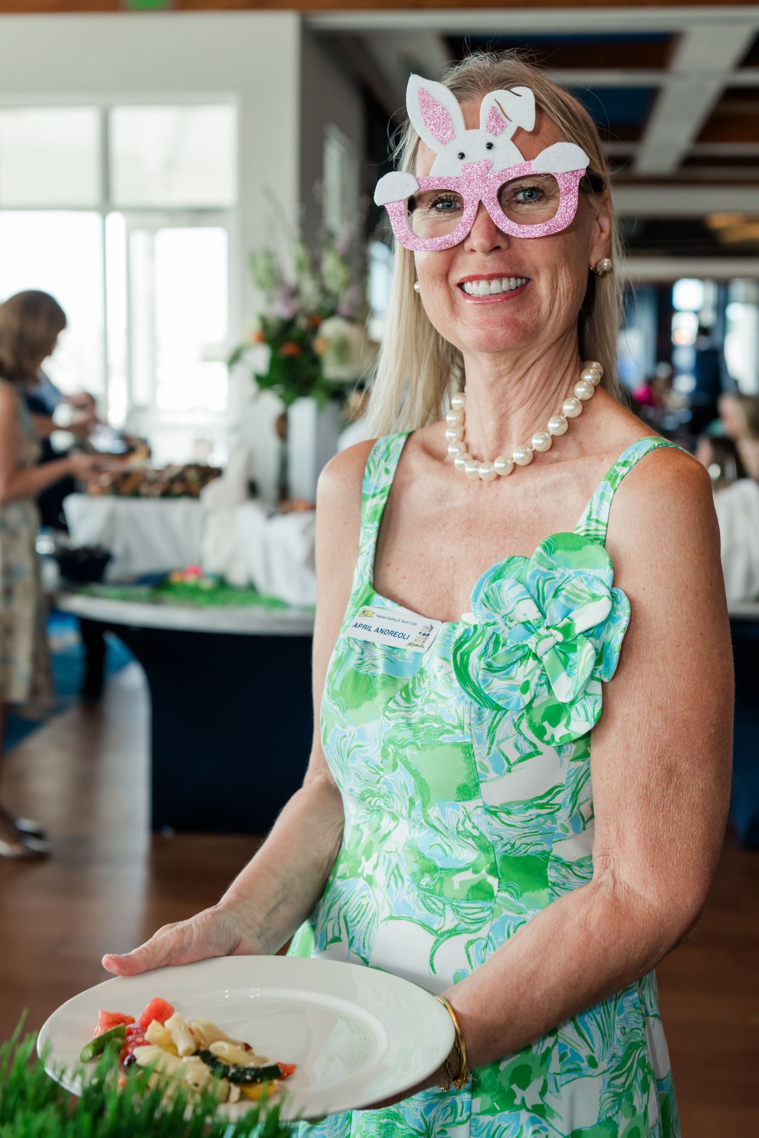 Woman in green floral dress wearing Easter bunny novelty glasses holds a plate at a festive spring event.